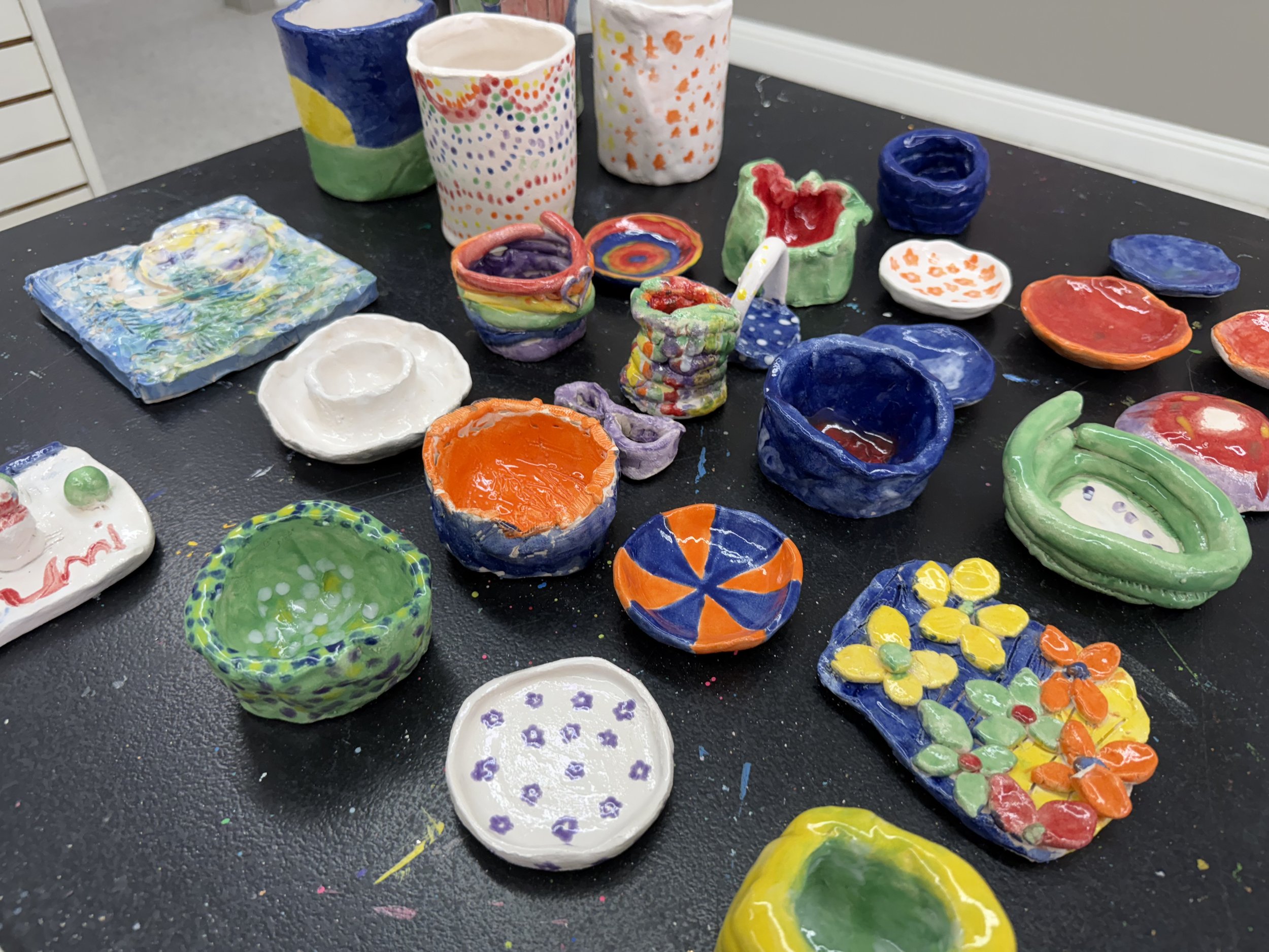 Colorful handmade ceramic bowls, cups, and dishes with various painted designs, arranged on a black table.