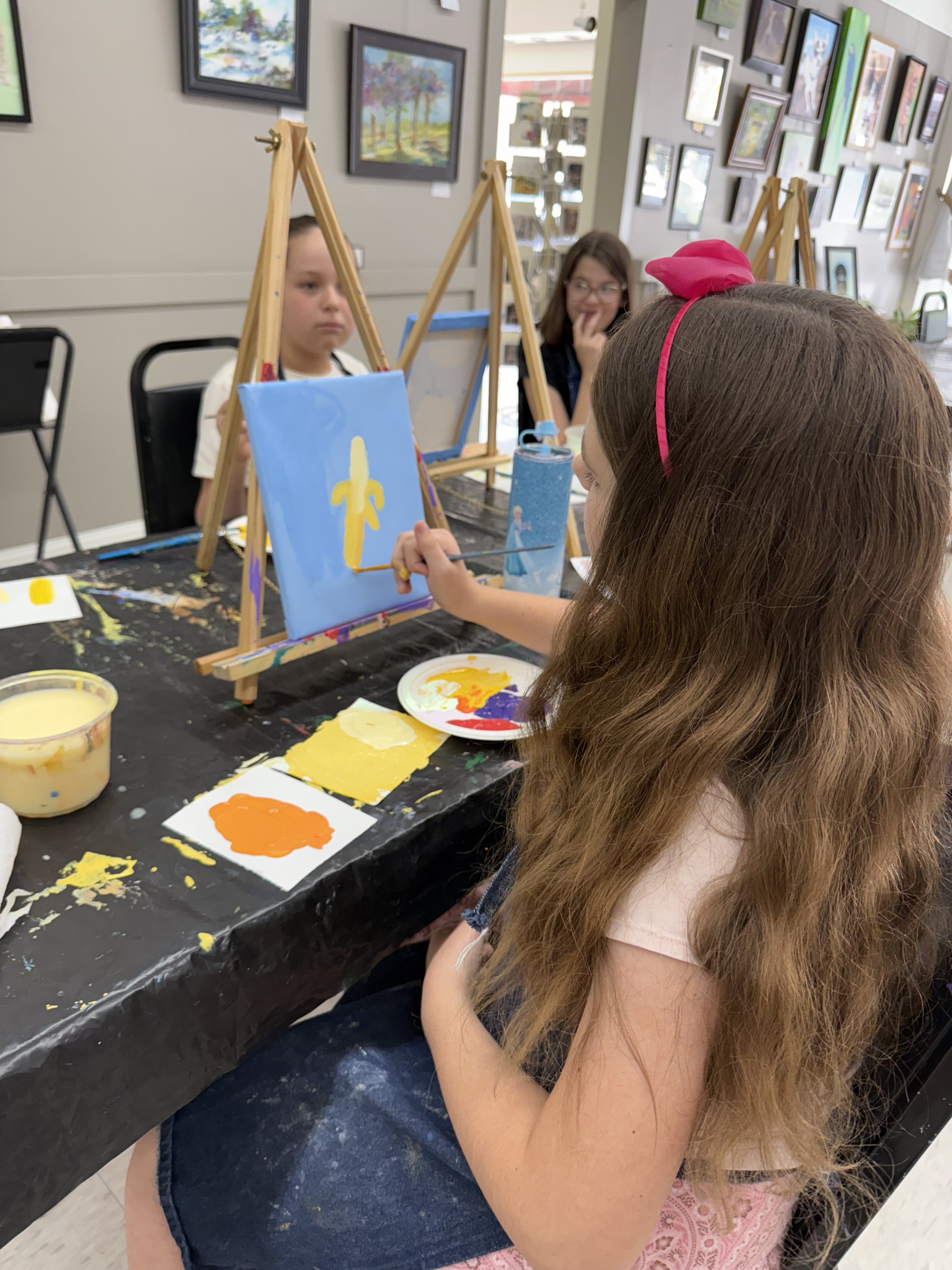 Children painting on small canvases at a classroom art table, with paintings and art supplies around.