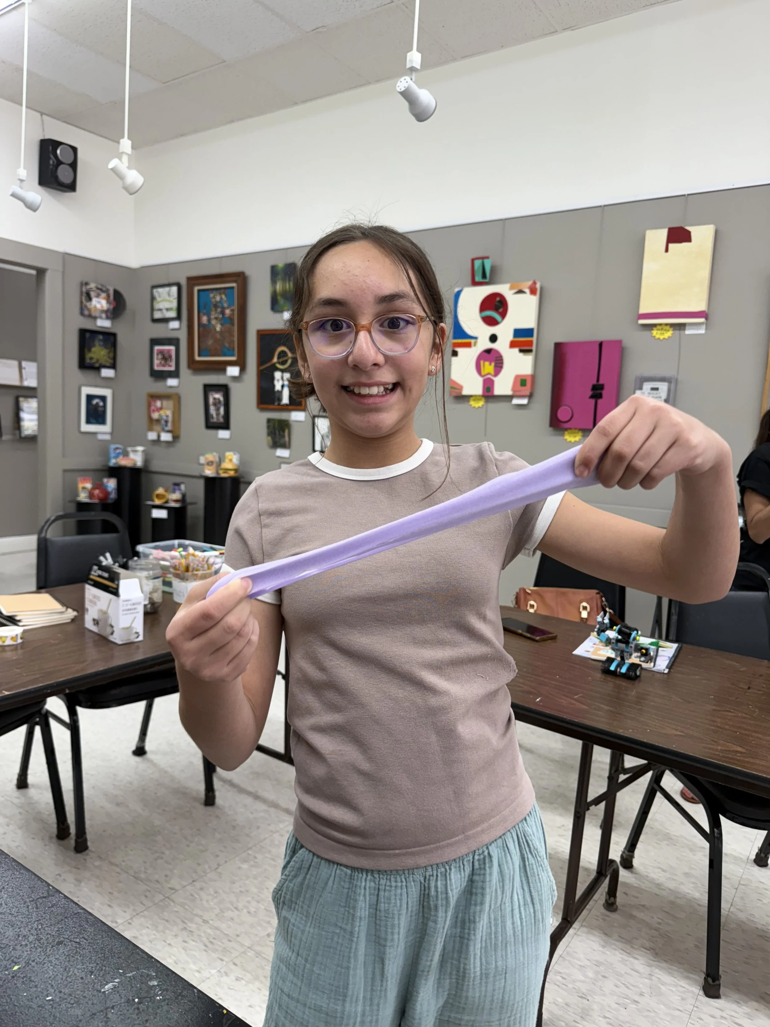 A young girl with glasses smiling and holding a long purple slime in an art classroom with artwork on the walls.