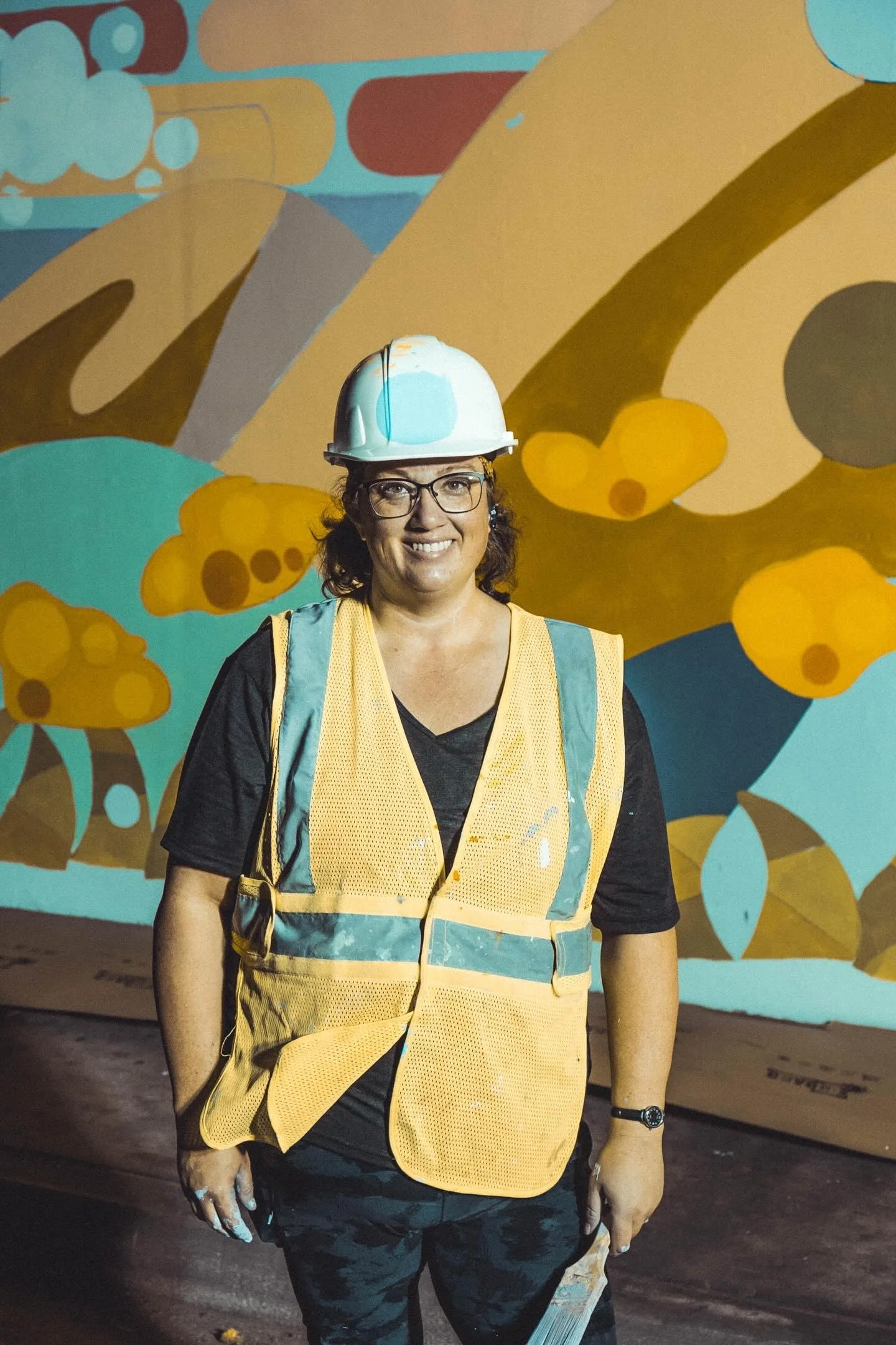 A woman with glasses wearing a white hard hat, yellow safety vest, and black shirt stands in front of a colorful mural with abstract shapes. She is smiling and holding a tool.