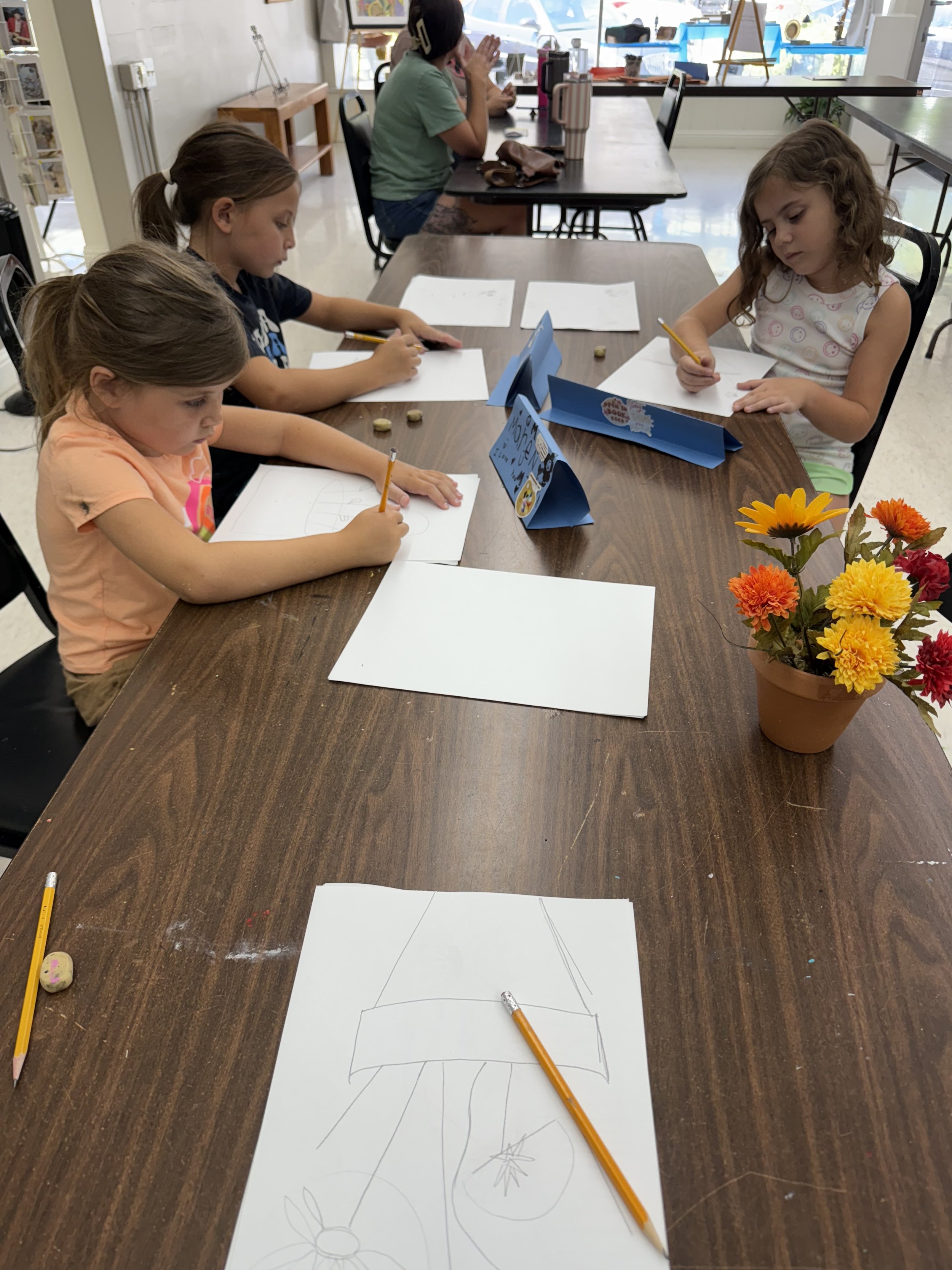 Four young girls sitting at a wooden table drawing on white sheets of paper with pencils. There are erasers and a small bouquet of colorful flowers on the table. In the background, two women are sitting at another table, facing the window and talking