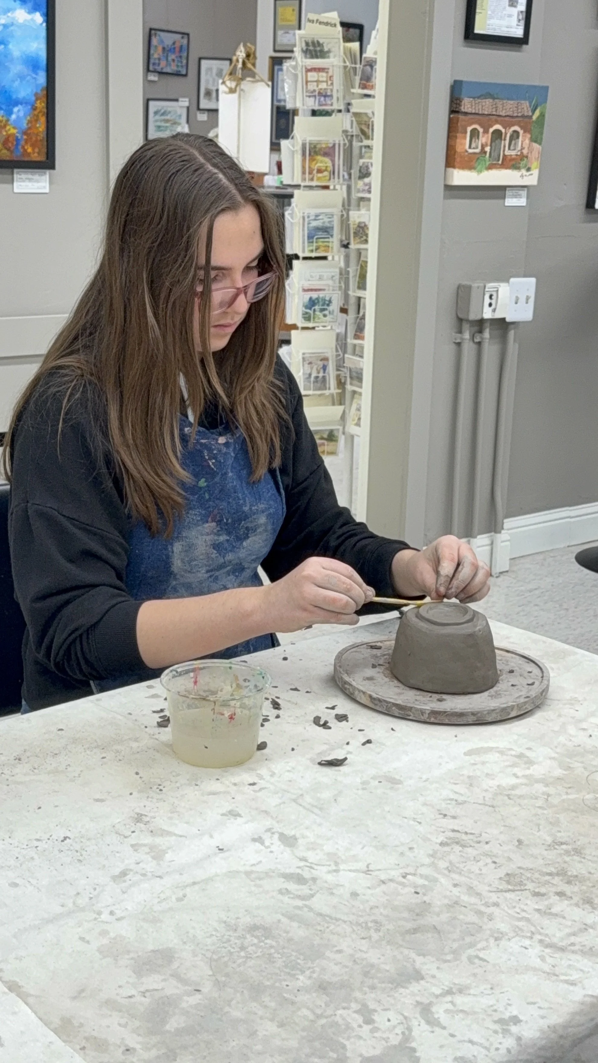 A young woman with long brown hair and glasses working on a pottery project, shaping a clay piece on a turntable in an art studio. The studio has artwork on the walls and art supplies on a table.