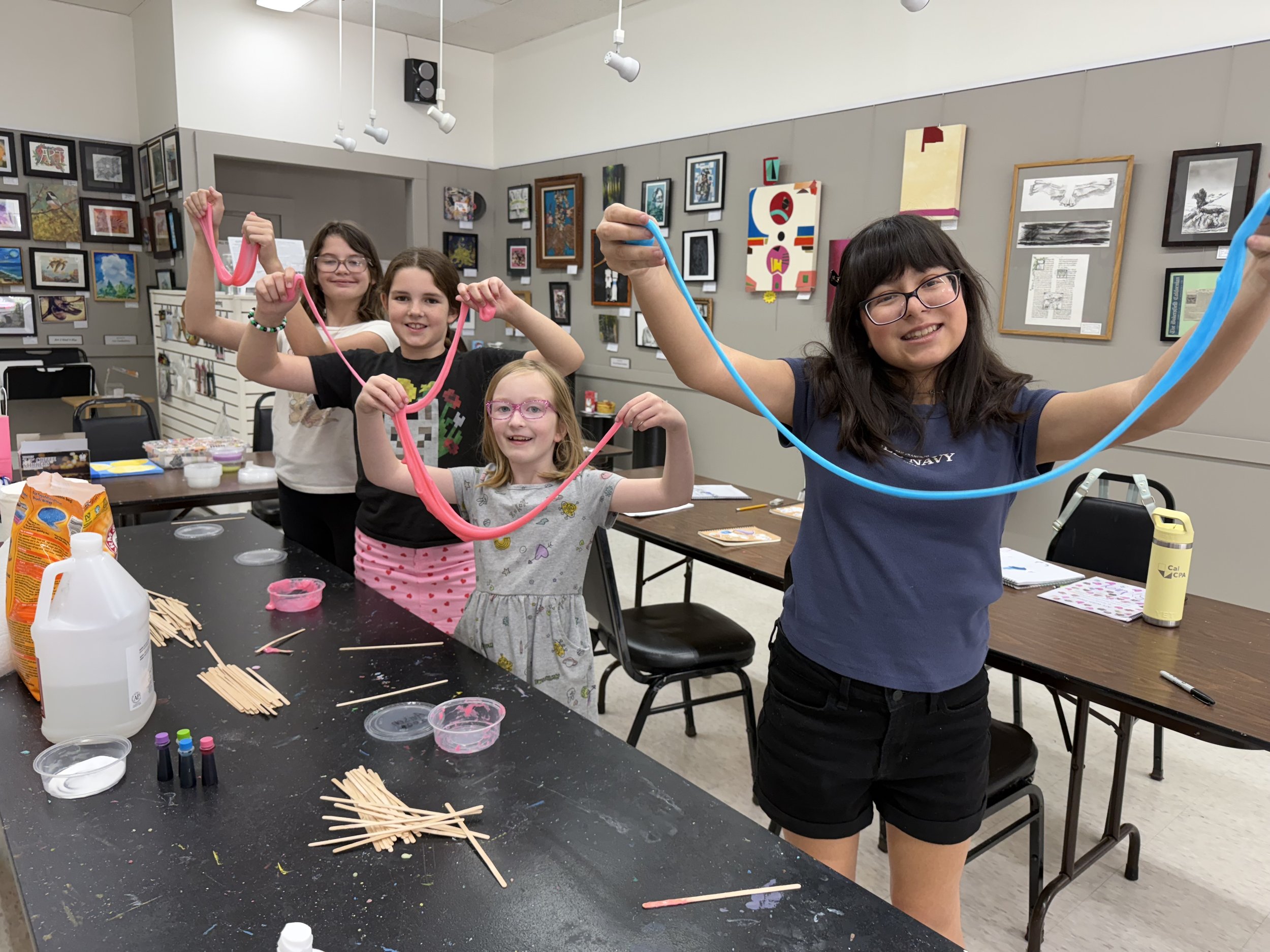 Group of five smiling children holding colorful slime strings in art room with artwork on walls and table with art supplies.