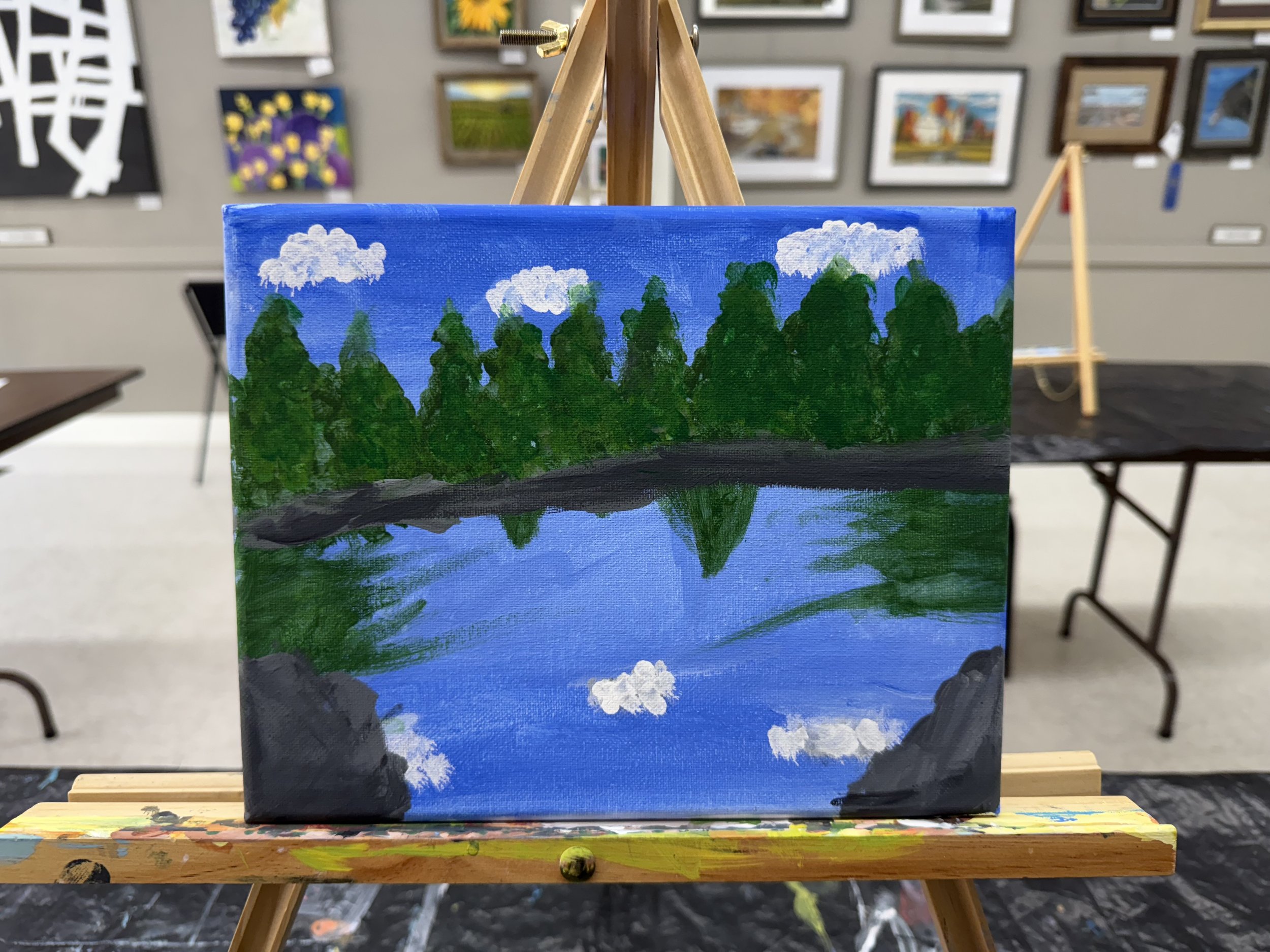 A landscape painting of a lake reflected with trees, clouds, and a blue sky, created with acrylic or oil paints on canvas, displayed on an easel in an art gallery or classroom.