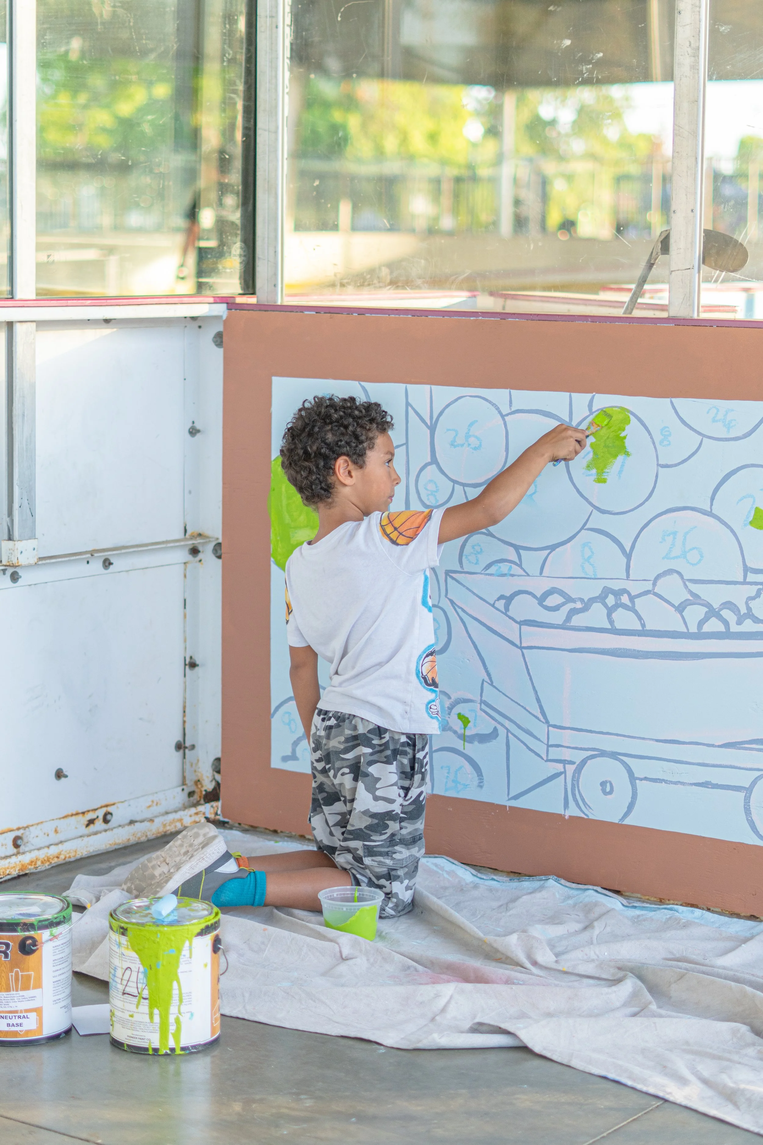 A young boy kneeling on the ground, painting a mural on a large board, with paint cans and brushes around him. The mural depicts a basket of apples and numbered circles, possibly for an educational activity.