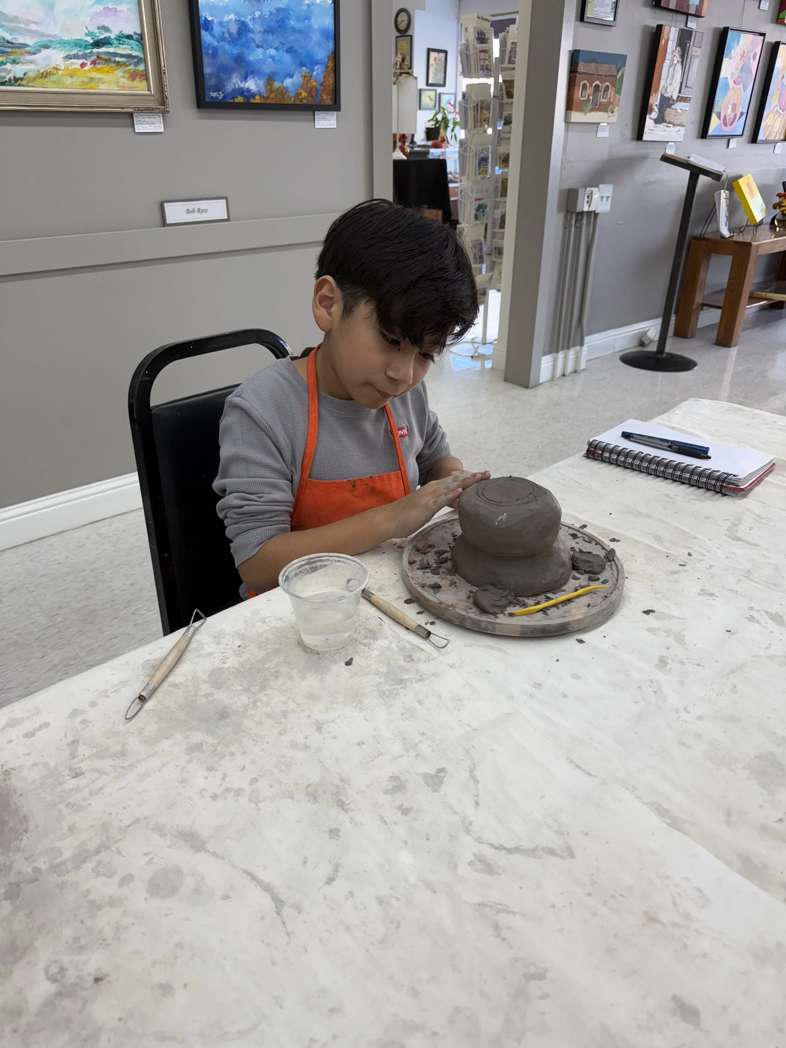 A young boy with dark hair wearing a gray shirt and orange apron is sitting at a table in an art gallery, working on a clay sculpture of a two-tiered cake. There are art paintings hanging on the walls behind him. On the table, there are pottery tools, a small cup of water, and a notebook with a pen.