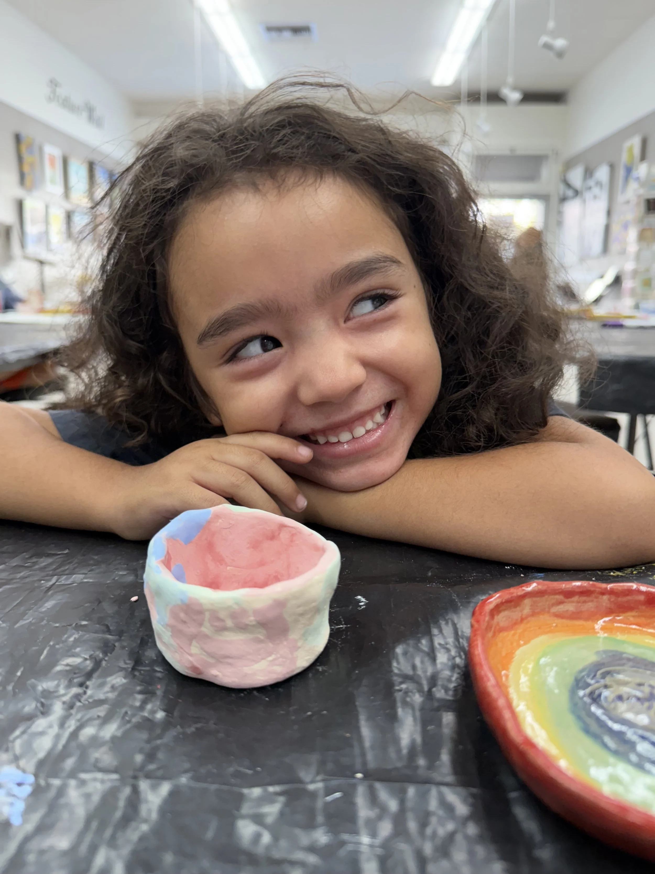 A young girl with curly hair and a big smile is resting her chin on her hand at a table covered with black paper. She is in an art classroom with colorful artwork on the walls behind her. On the table in front of her are a small, colorful, handmade pottery bowl and a rainbow-colored ceramic dish.