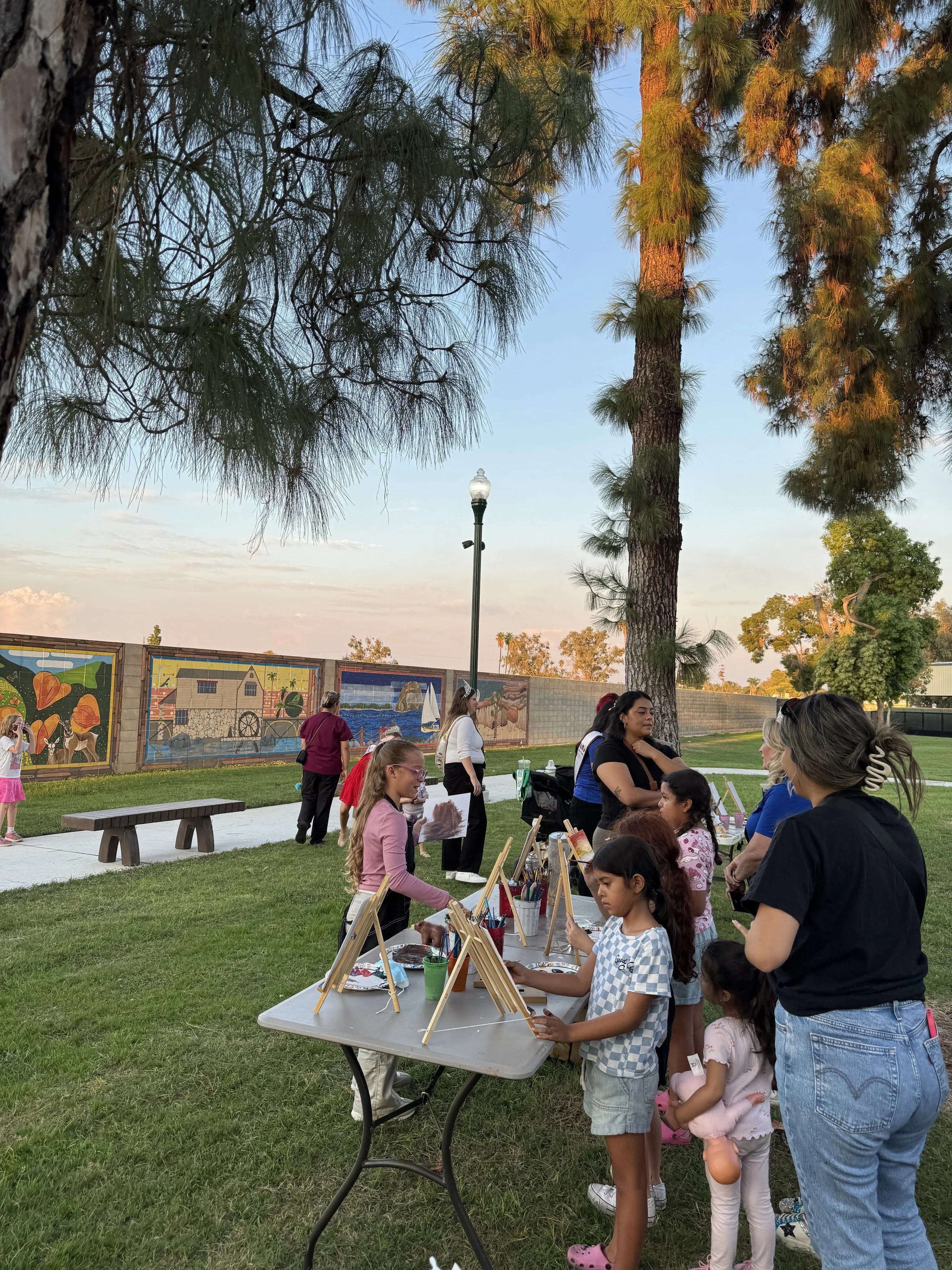 Children and adults participating in an outdoor painting activity at a park during sunset, with trees and murals in the background.