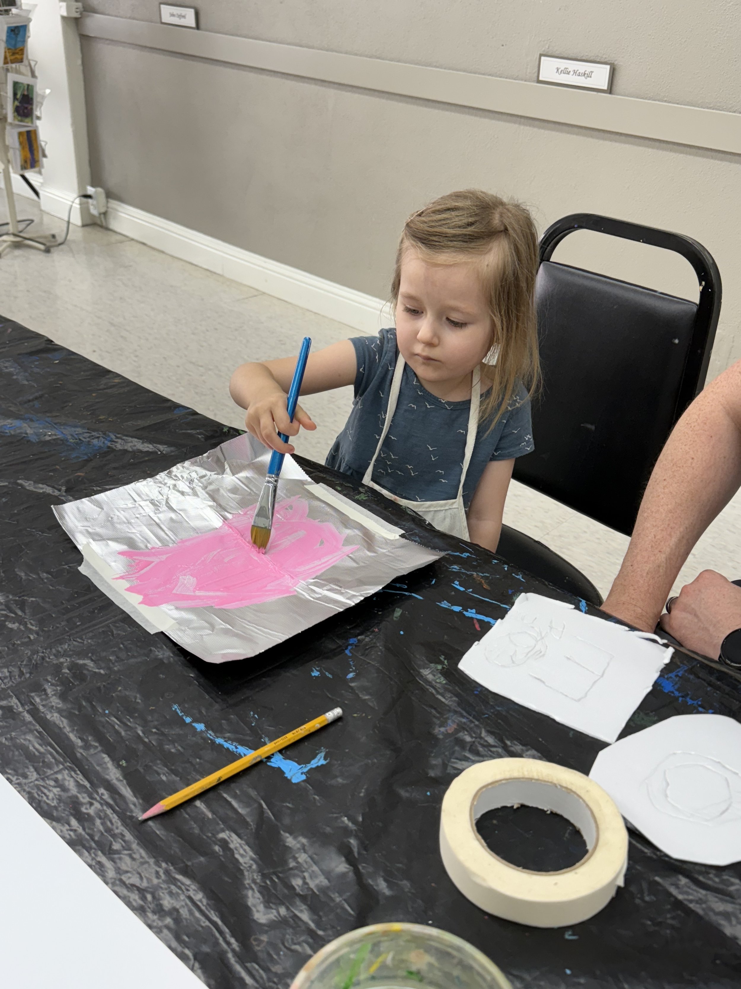 A young girl with a blue shirt and apron painting pink on a piece of aluminum foil with a brush, sitting at a table covered with black plastic and art supplies, with a rolled piece of tape and a pencil on the table.