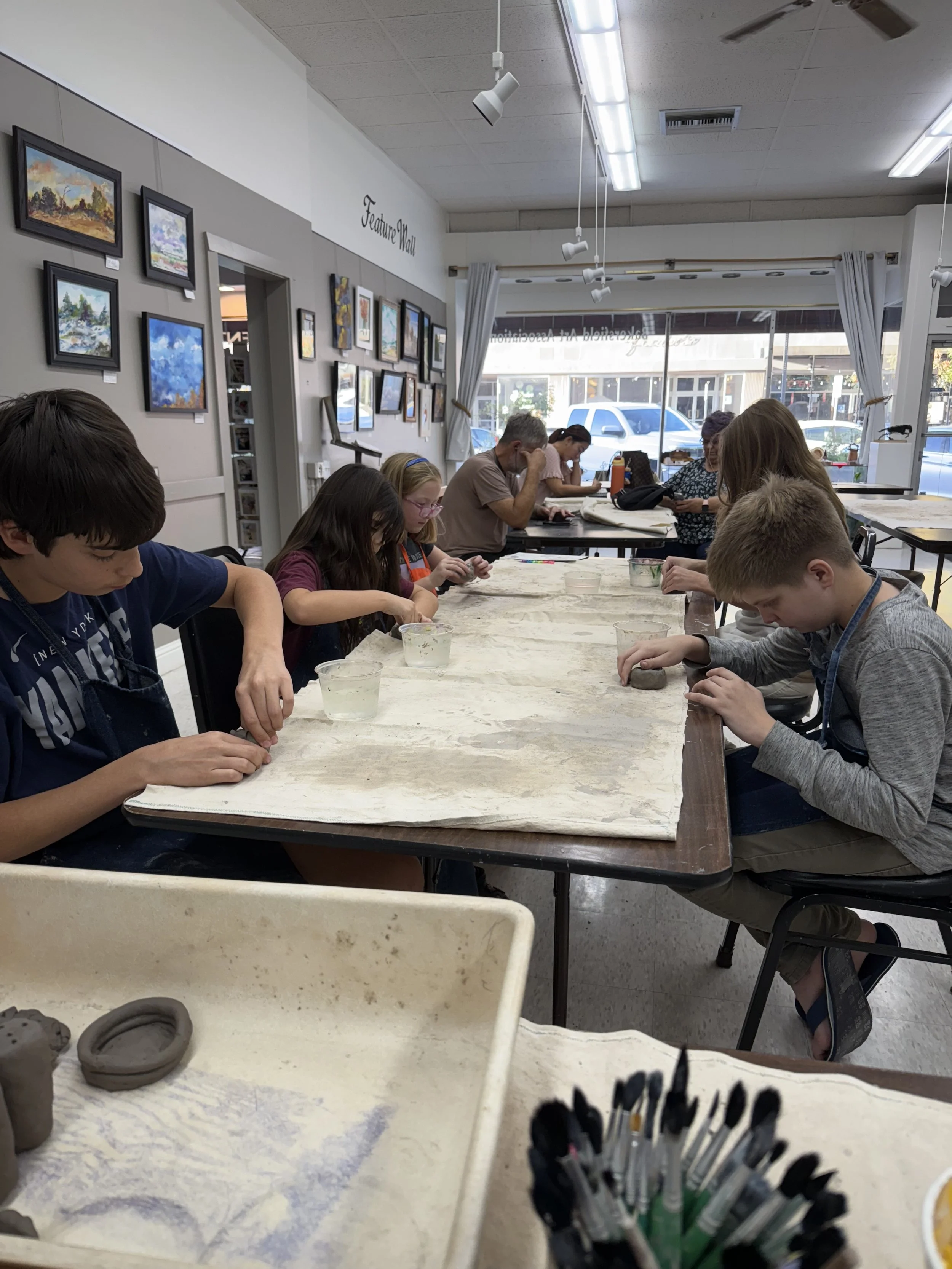 Children and adults are seated at a long table in an art studio, working on clay projects. The studio has framed artwork on the walls and large windows allowing natural light inside.