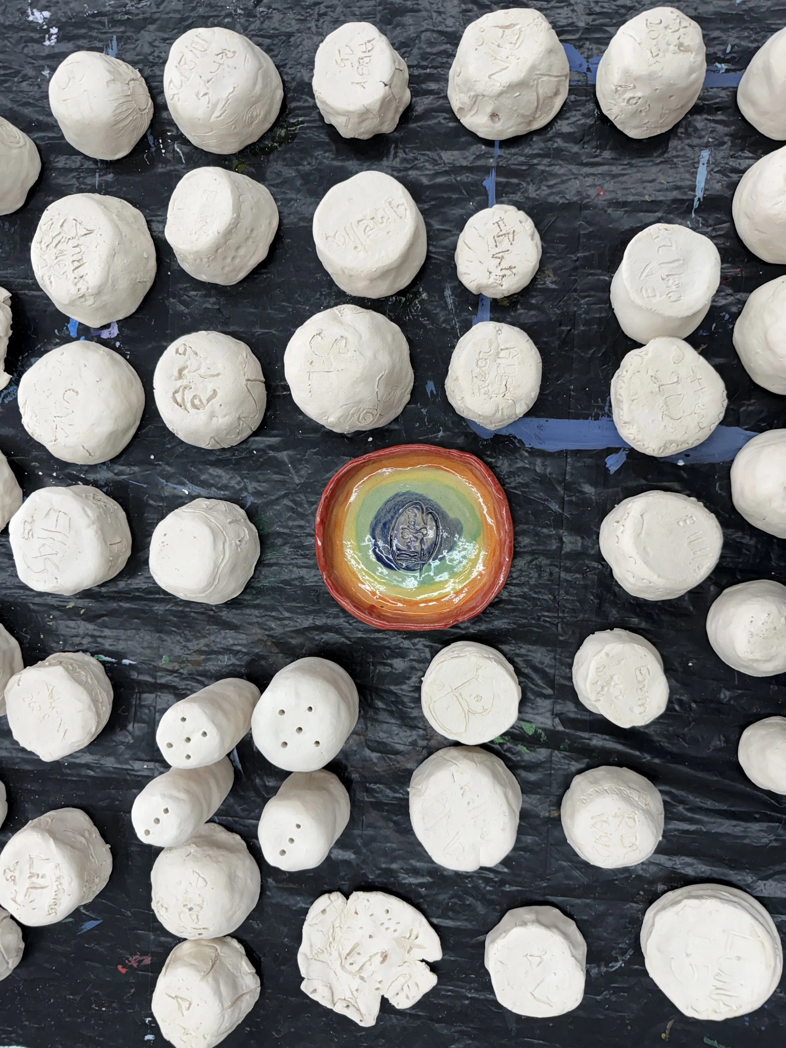 Collection of white clay or plaster balls with handwritten inscriptions in a grid pattern, centered on a black surface with a colorful round object with a rainbow pattern in the middle.