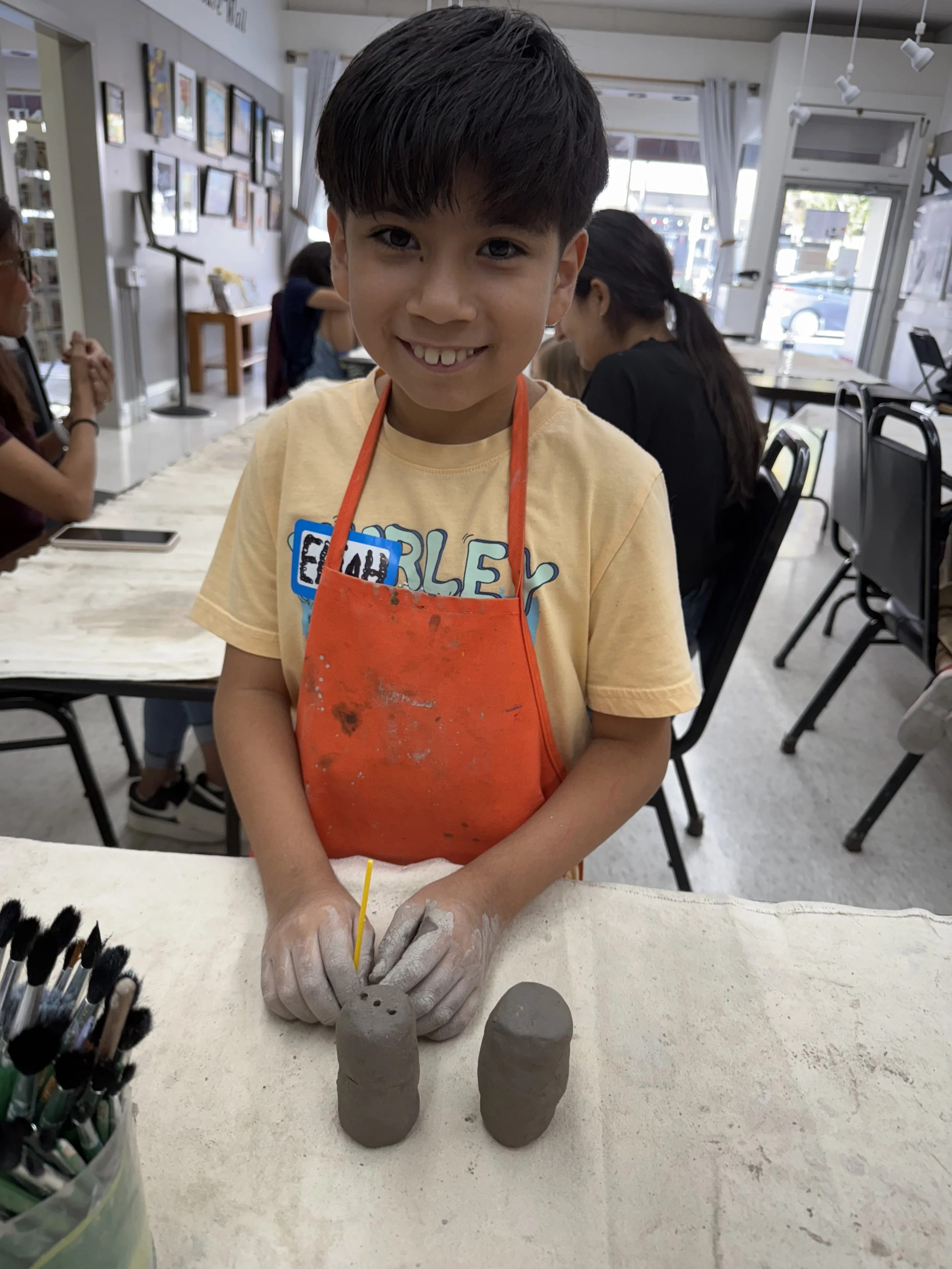 Young boy with a yellow shirt and orange apron smiling at the camera, standing at a table with two clay sculptures, while working on them with a small tool. In the background, other children and adults are seated at tables in a well-lit art classroom