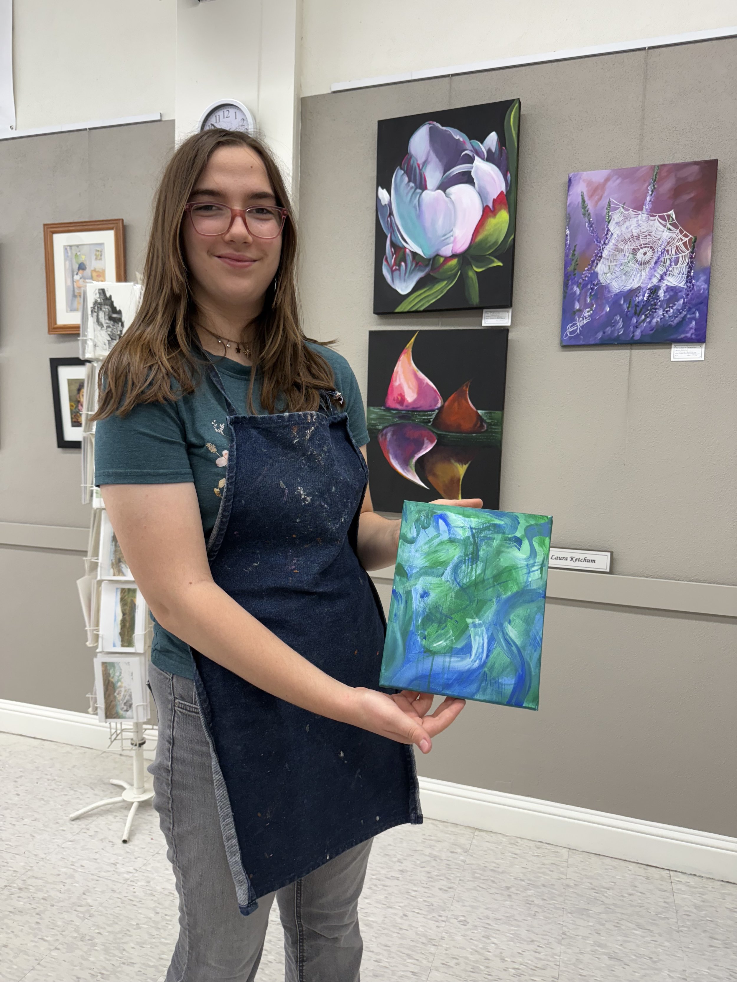 Young woman with glasses and a smirk, wearing a dark apron, holding a small abstract painting in a gallery or art exhibit. Behind her are various colorful paintings and framed artworks on a gray wall.