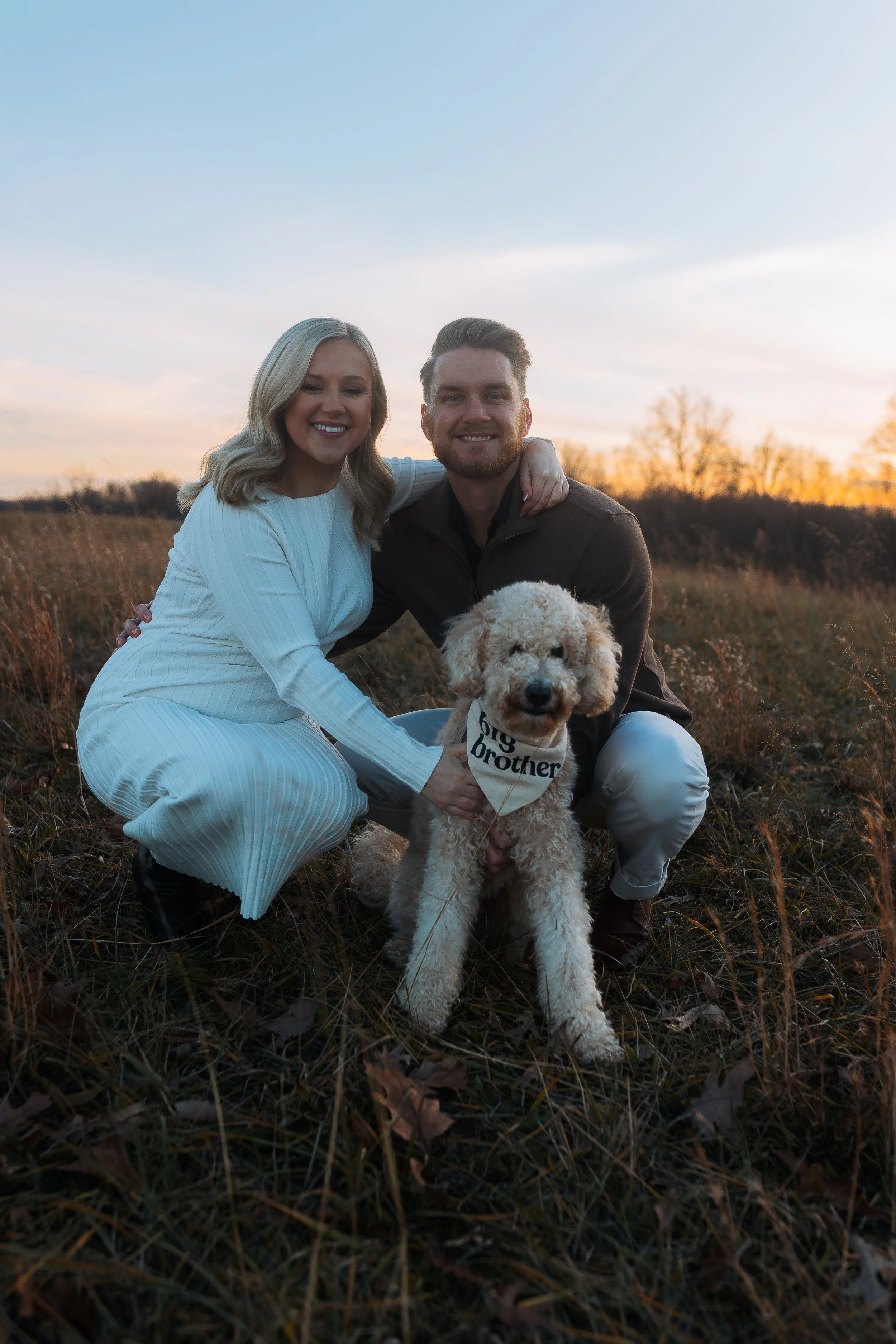 A couple in an open field at sunset, with a dog wearing a bandana that says 'Big Brother,' smiling and posing for the photo.