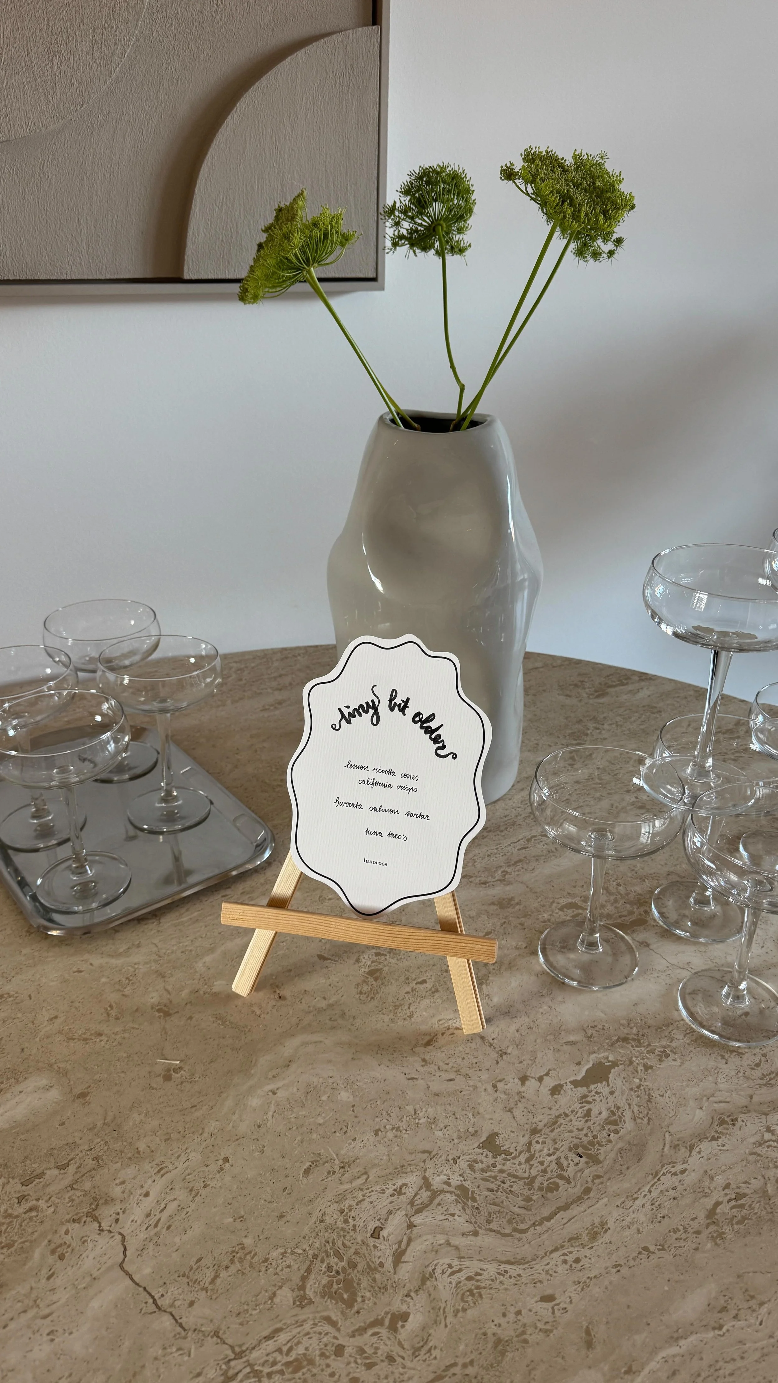 Een witte vaas met groene bloemen, een klein bordje op een houten ezel met een lijntekening en lege glazen op een marmeren tafel.