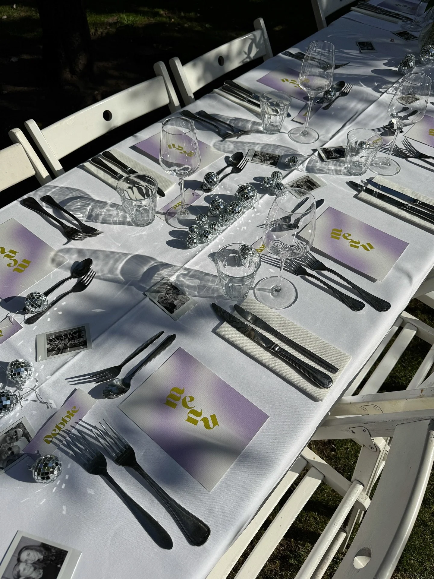 Een feestelijk gedekte tafel met een wit tafelkleed, wijnglazen, bestek, zwarte servetten, paarse en gele menu’s en kleine geruite foto‑ornamenten, buiten opgesteld in zonlicht en schaduw voor catering.
