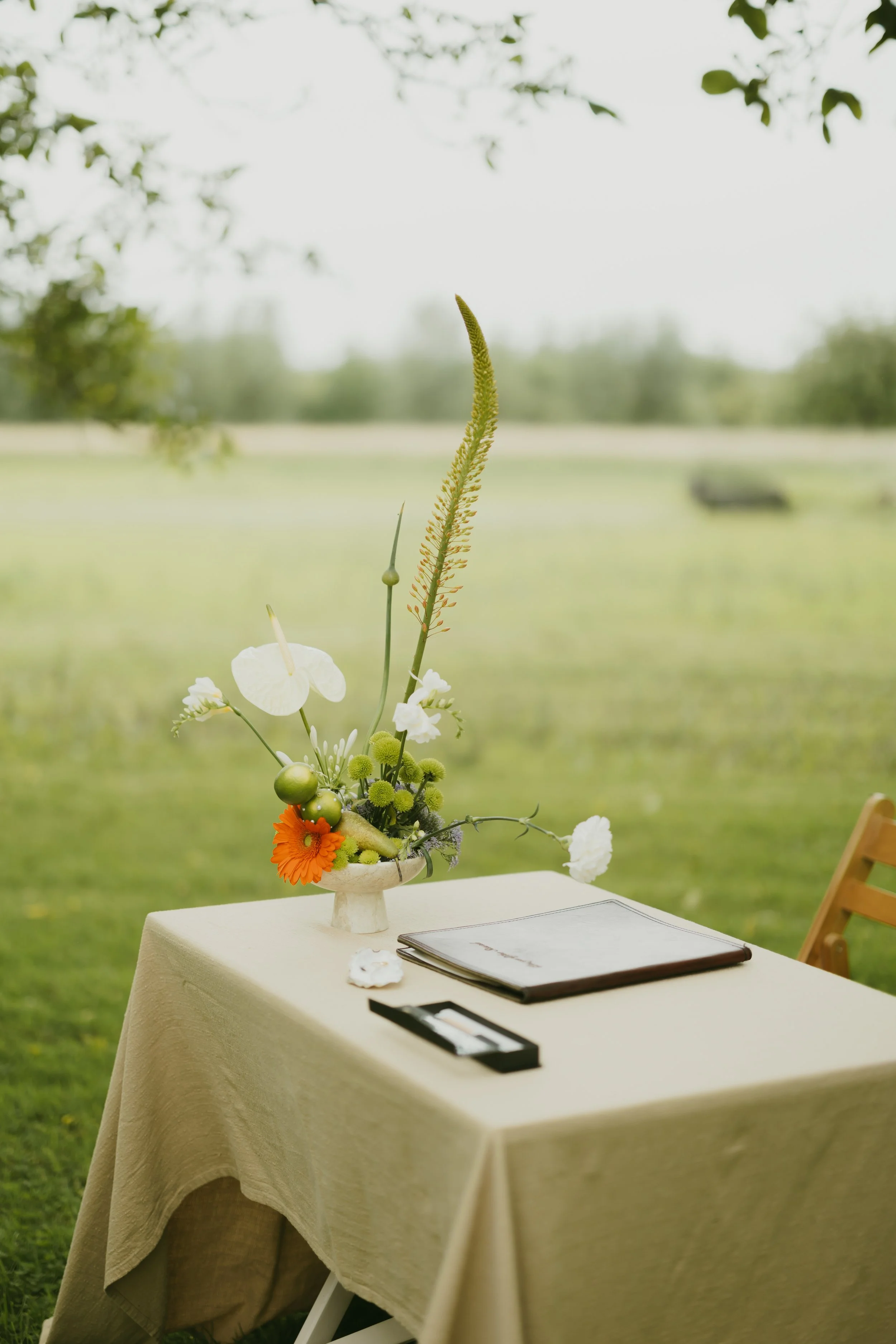 Trouw tafel met een groot bloemstuk, akteboekjes en een bril, opgesteld buiten op een groene grasmat.