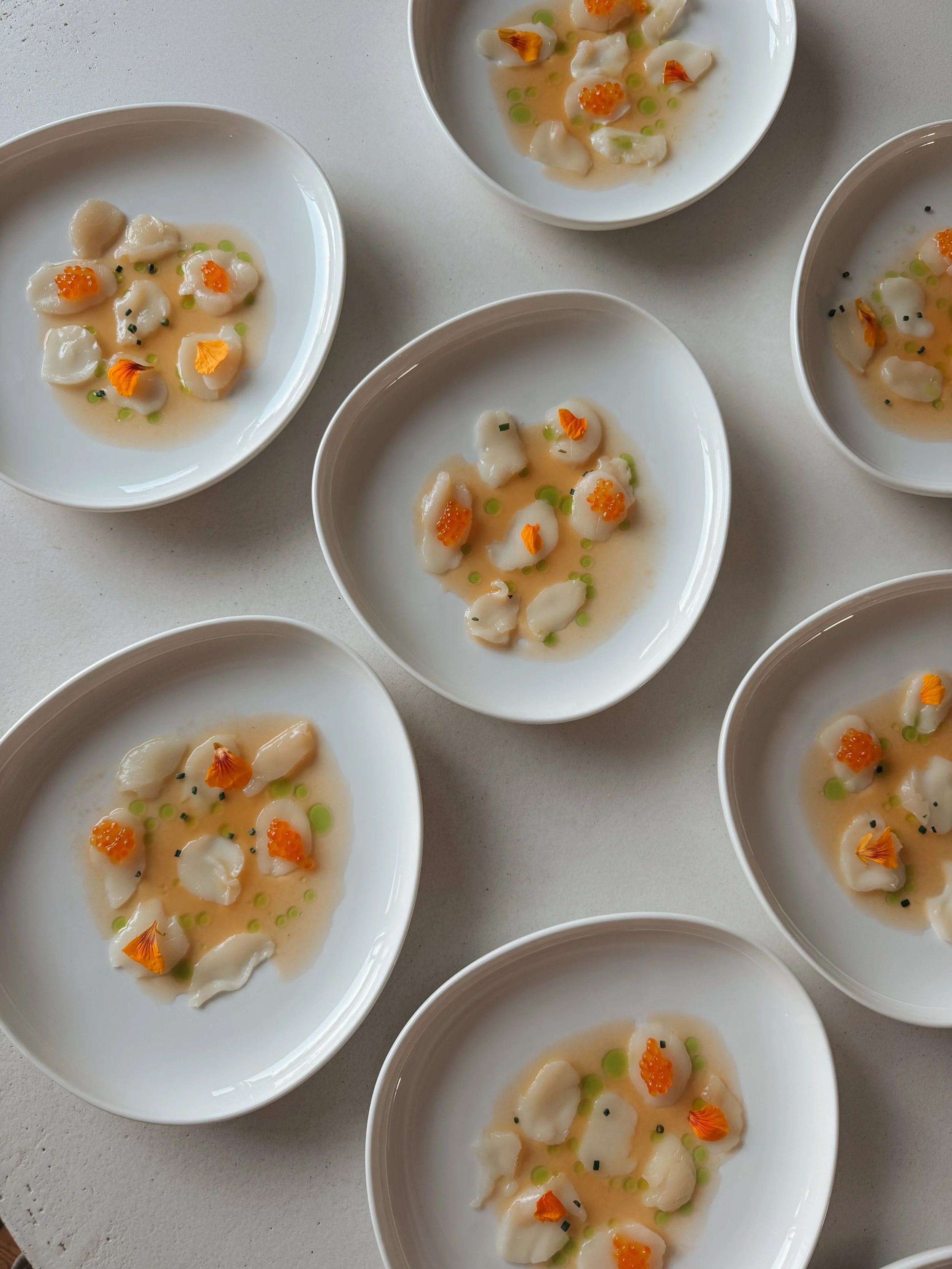 Meerdere witte schalen met kleine stukjes zeevruchten, gegarneerd met oranje en groene toppings, op een witte ondergrond voor een catering.