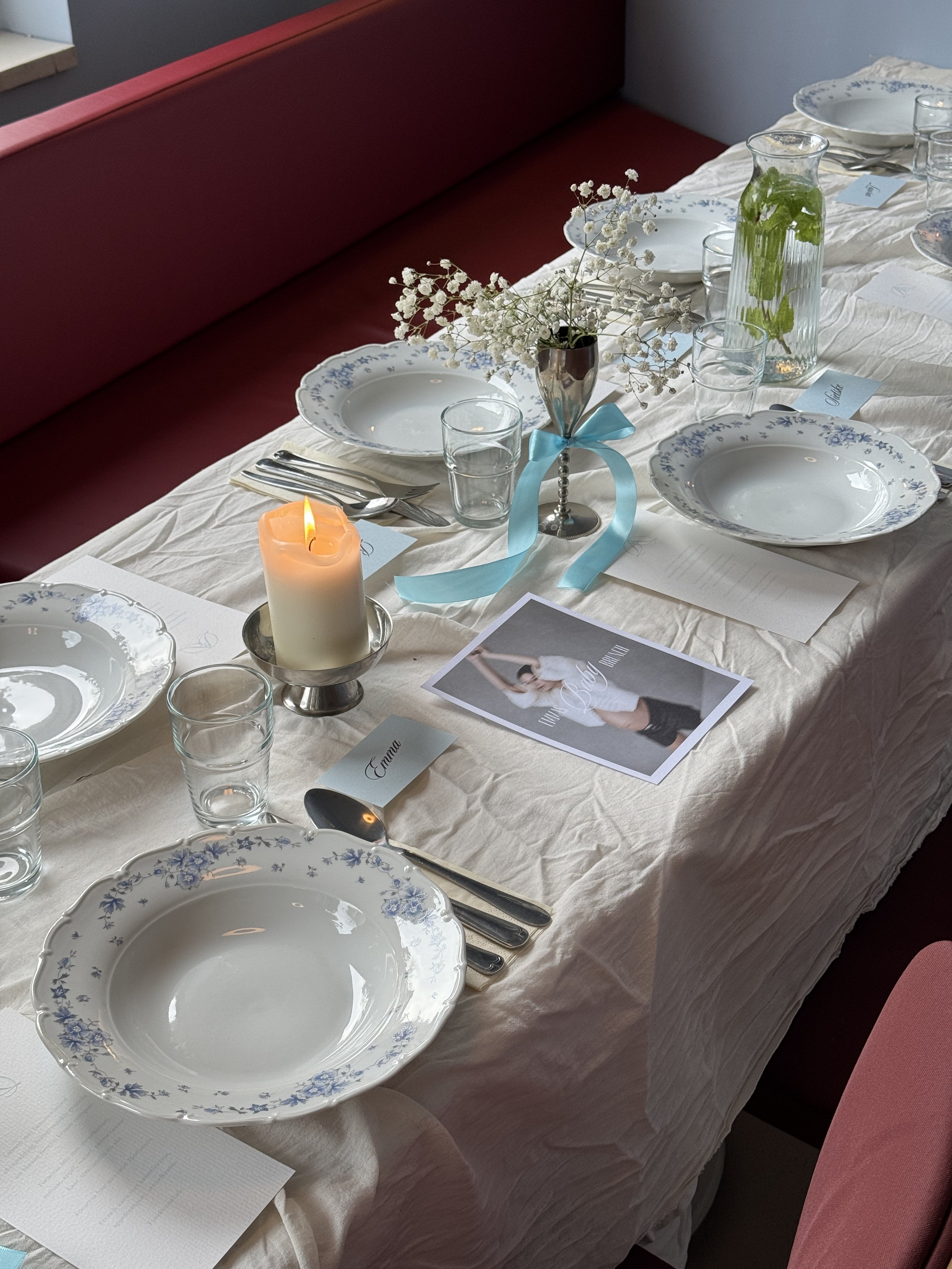 Een gedekte dinertafel voor een formele maaltijd, met wit‑blauw bloemmotief servies, heldere glazen, een witte kaars, een klein bloemstukje met gipskruid en naamkaartjes. Op de tafel ligt een foto van een vrouw in wit en zwart, en op één naamkaartje.