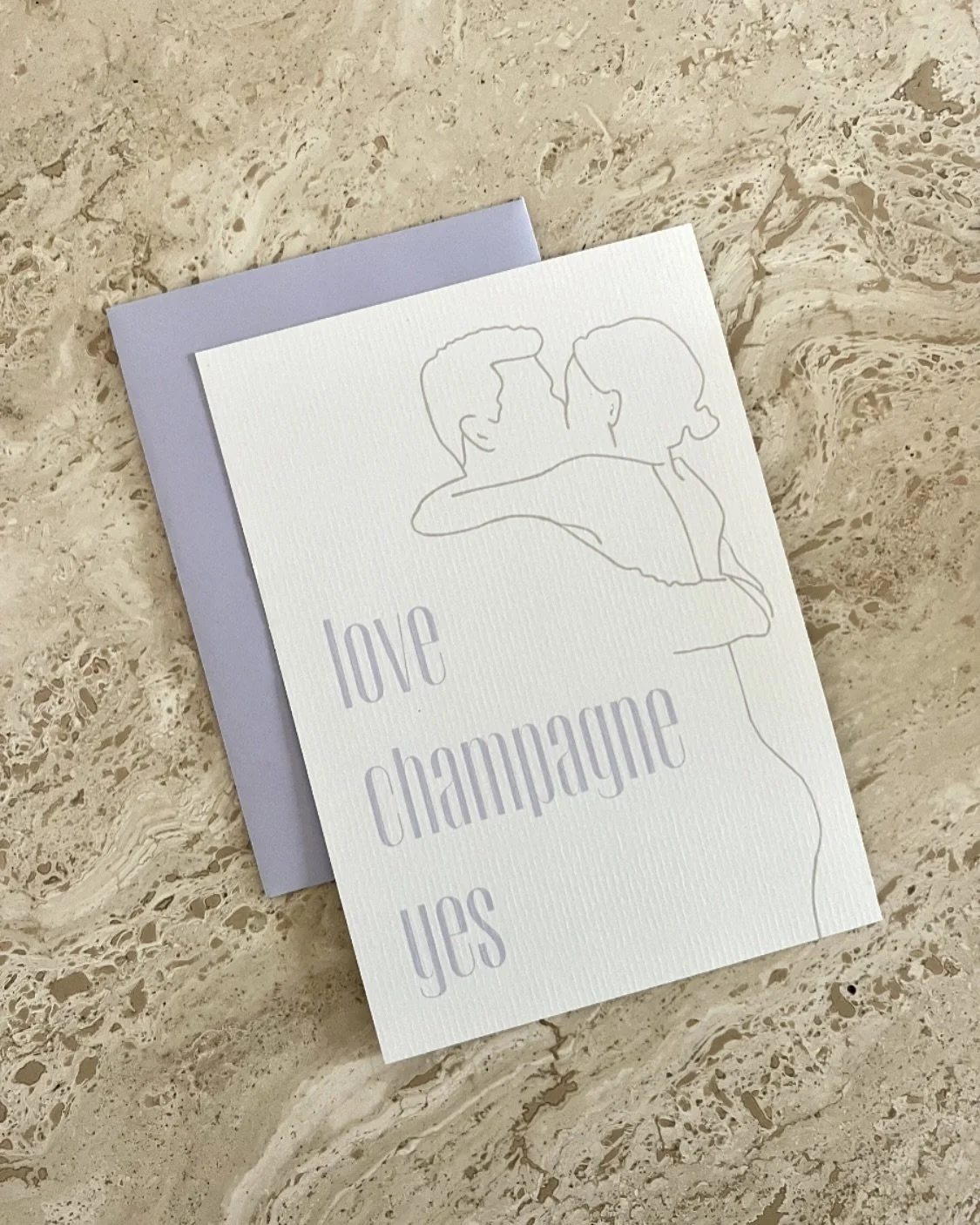 Een trouwkaart met een minimalistische lijntekening van twee omhelzende personen en de woorden ‘love champagne yes’, geplaatst op een marmeren oppervlak.