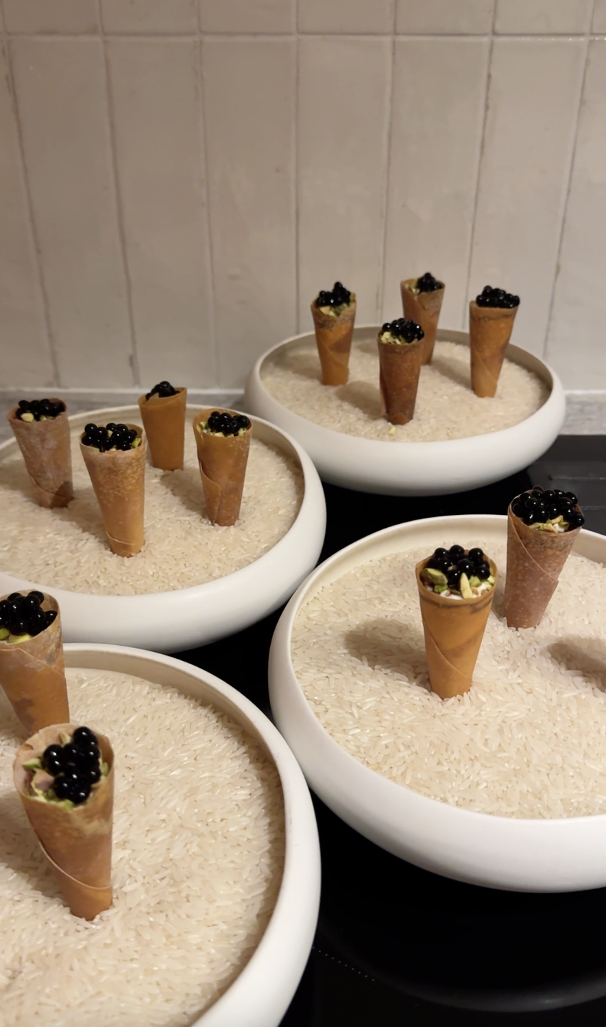 Een gedekte eettafel met twee witte kaarsen, twee wijnglazen, een waterglas, een kom met een witte romige schotel, een lege kom en groene plaatskaartjes met tekst, in zonlicht.