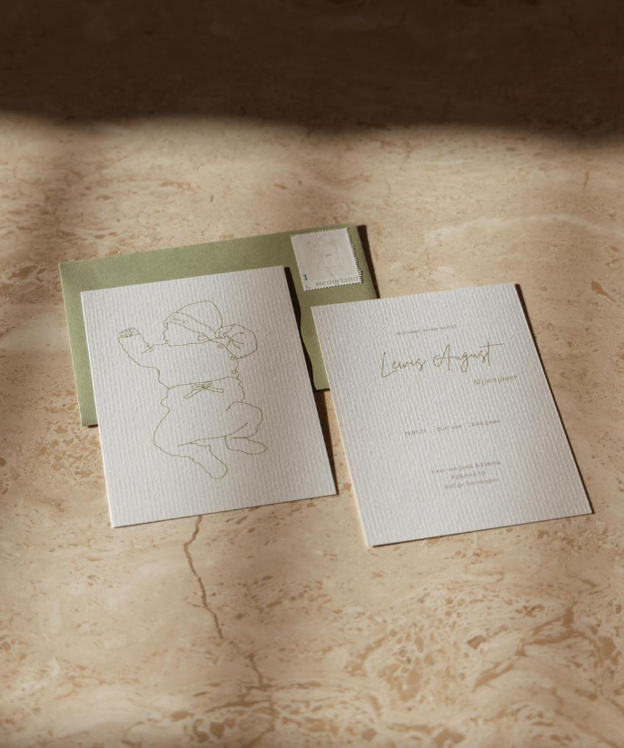 Een collectie stationery op beige marmer, waaronder een wenskaart met een lijntekening van een kind in een jas met capuchon, een envelop en een uitnodigingskaart met gouden cursieve tekst ‘Lewis August’.