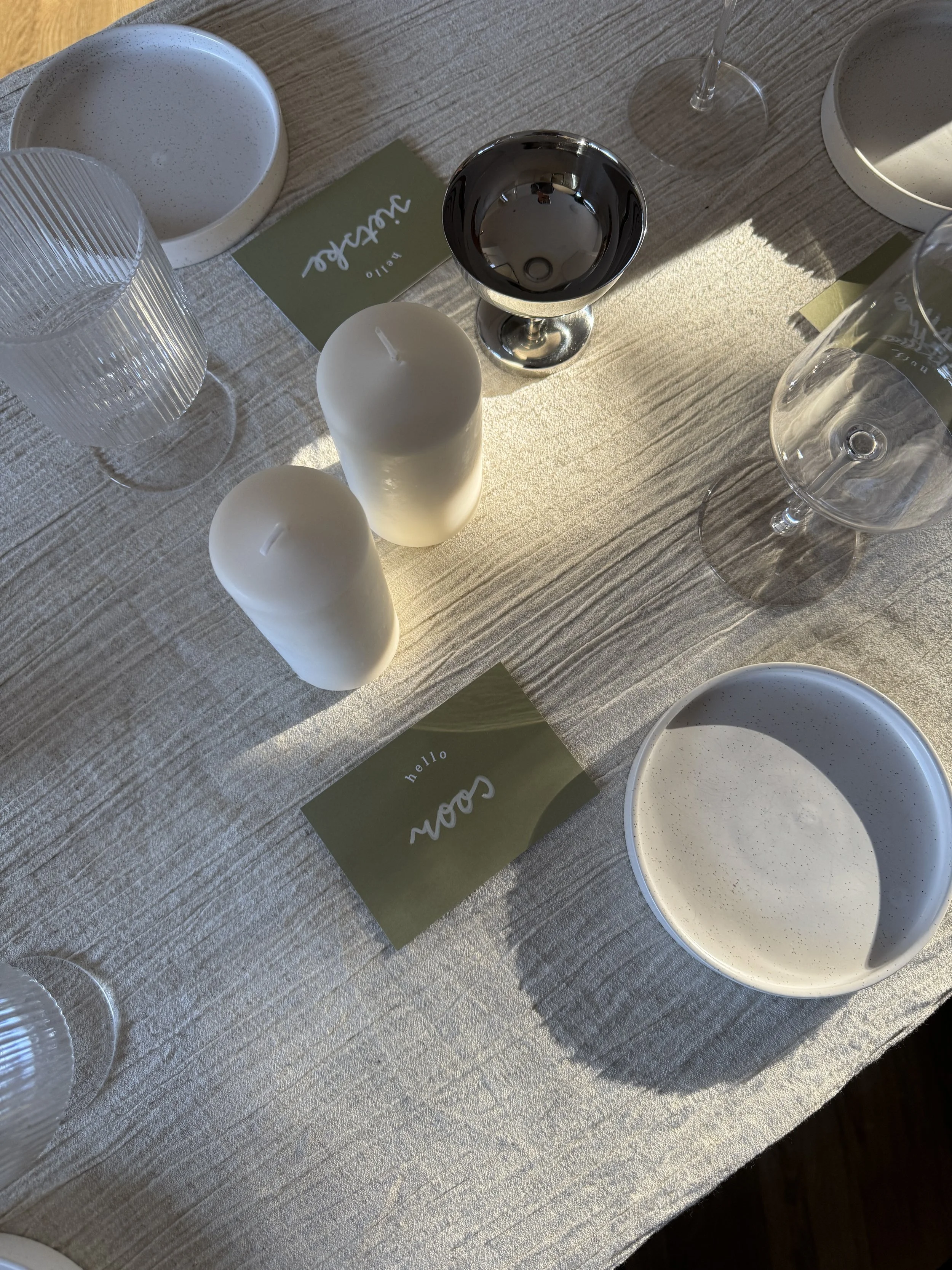 Een gedekte eettafel met twee witte kaarsen, twee wijnglazen, een waterglas, een kom met een witte romige schotel, een lege kom en groene plaatskaartjes met tekst, in zonlicht voor een catering opdracht.