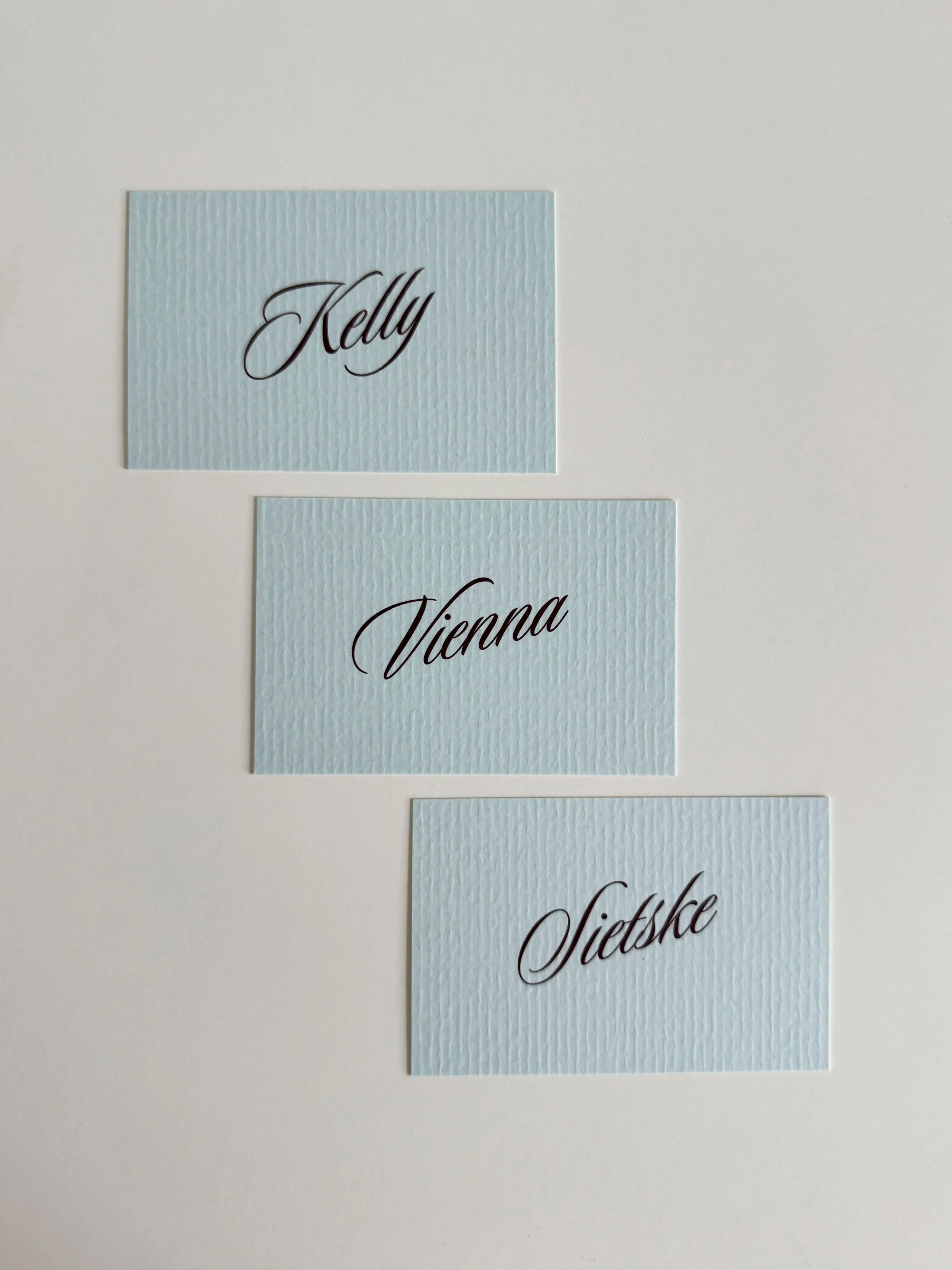 Drie lichtblauwe naamkaartjes met textuur en zwarte sierlijke tekst, verticaal gerangschikt, elk met een naam: Kelly, Vienna, Sietske.