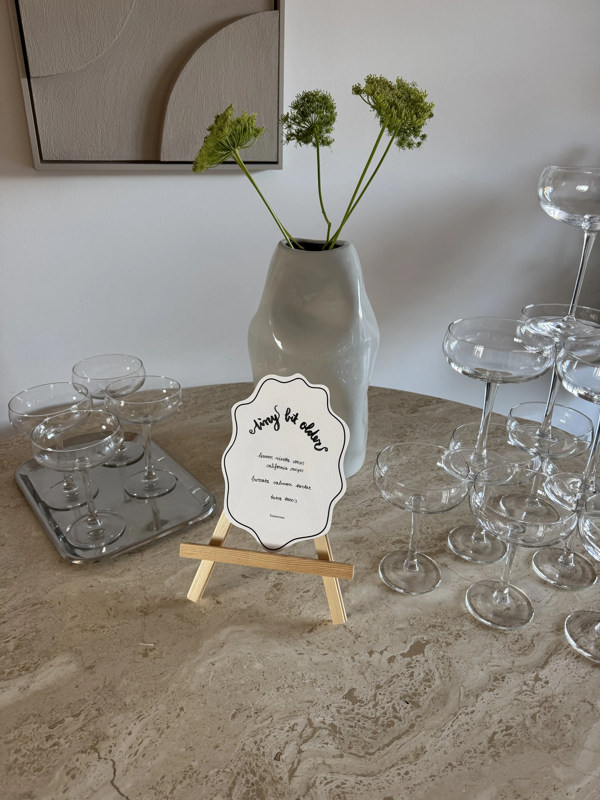 Een witte keramische vaas met groene bloemen op een beige marmeren tafel, omringd door lege glazen voor hapjes en cocktails, met een klein menu op een houten standaard en muurkunst op de achtergrond.