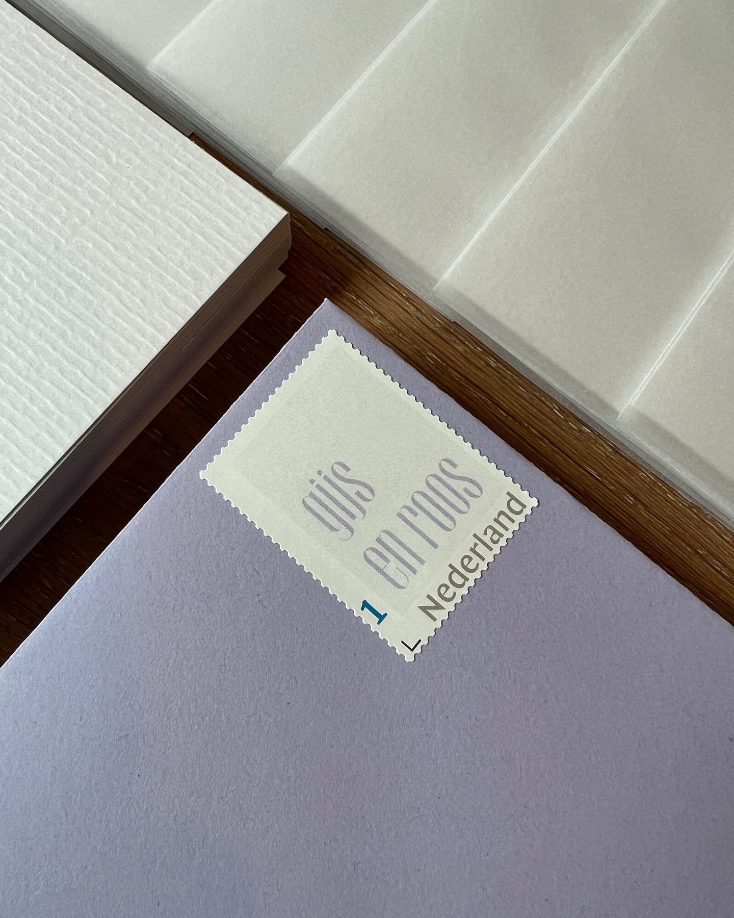 Close‑up van een postzegel met de woorden ‘LOVE’ en ‘Nederland’ op een paars stuk papier, naast een stapel wit papier en een witte plastic lade met rechthoekige compartimenten.