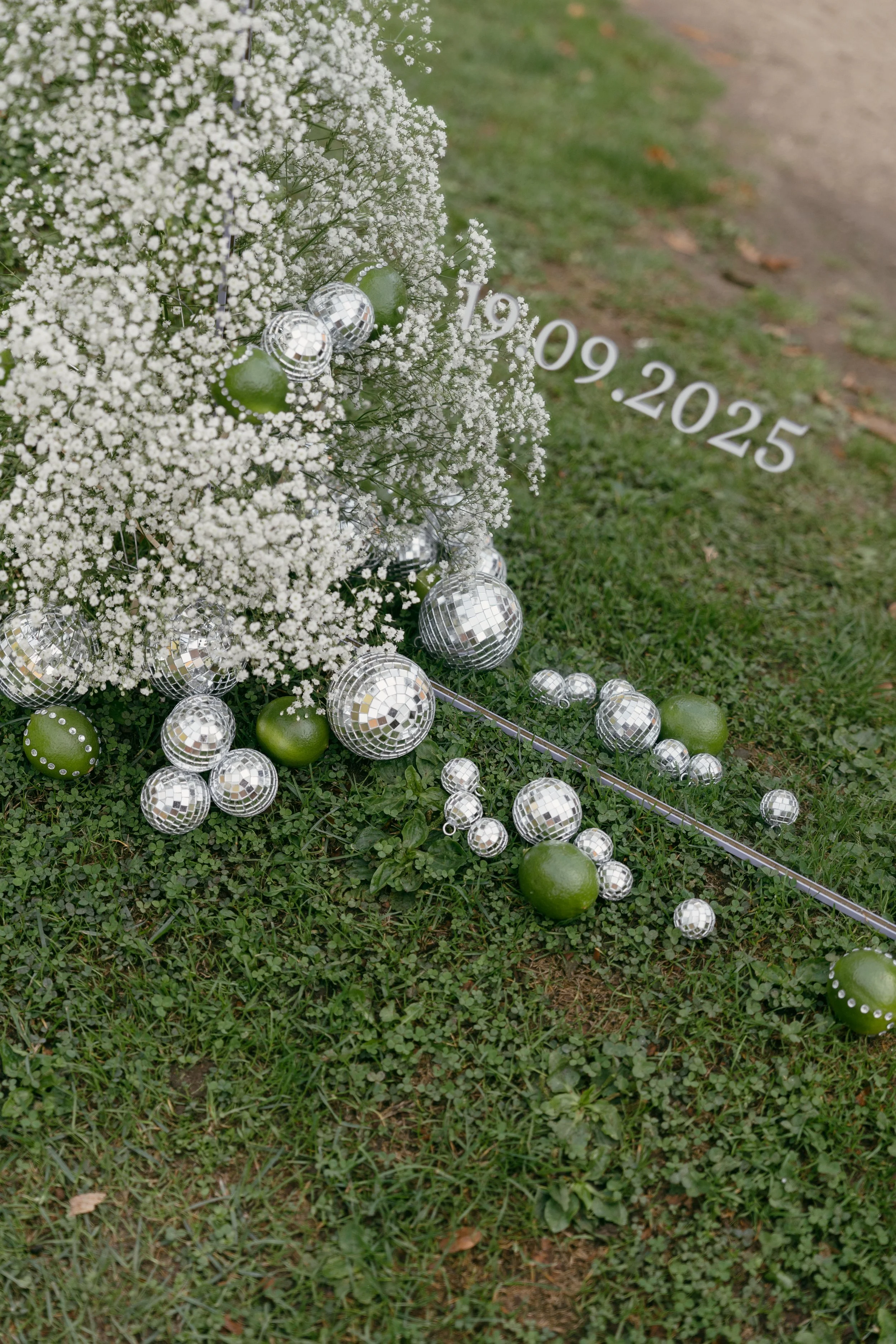 Decoratieve opstelling bruiloft met kleine witte bloemen, welkomst spiegel, hangende zilveren discoballen en groene ornamenten op het gras, met een bord waarop de trouwdatum staat weergegeven.