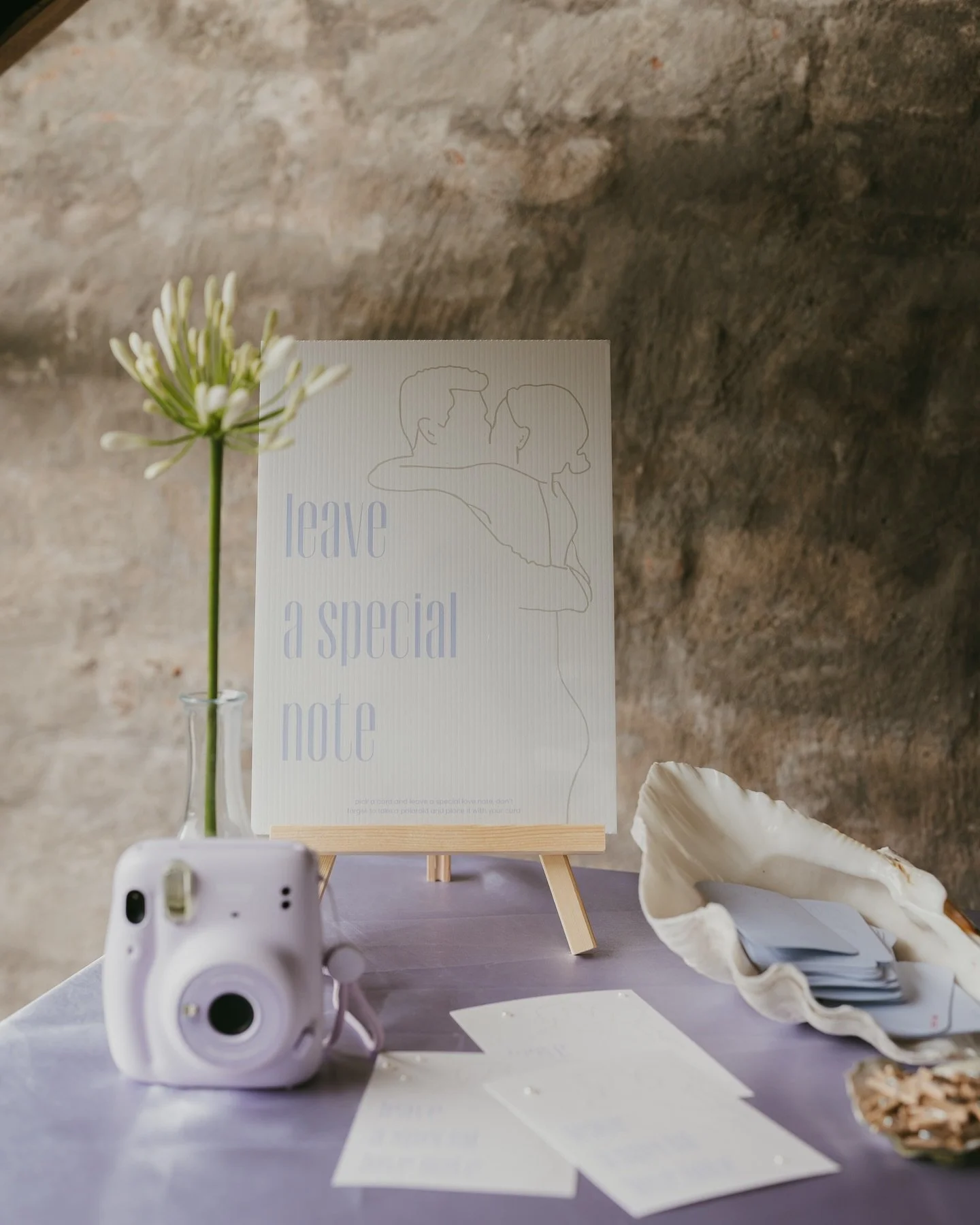 Tafeldecoratie voor een bruiloft met een klein vaasje met een bloem, een witte camera, een uitnodigingskaart, een kleine ezel met een notitie, schelpen en papieren, geplaatst tegen een ruwe stenen muur.