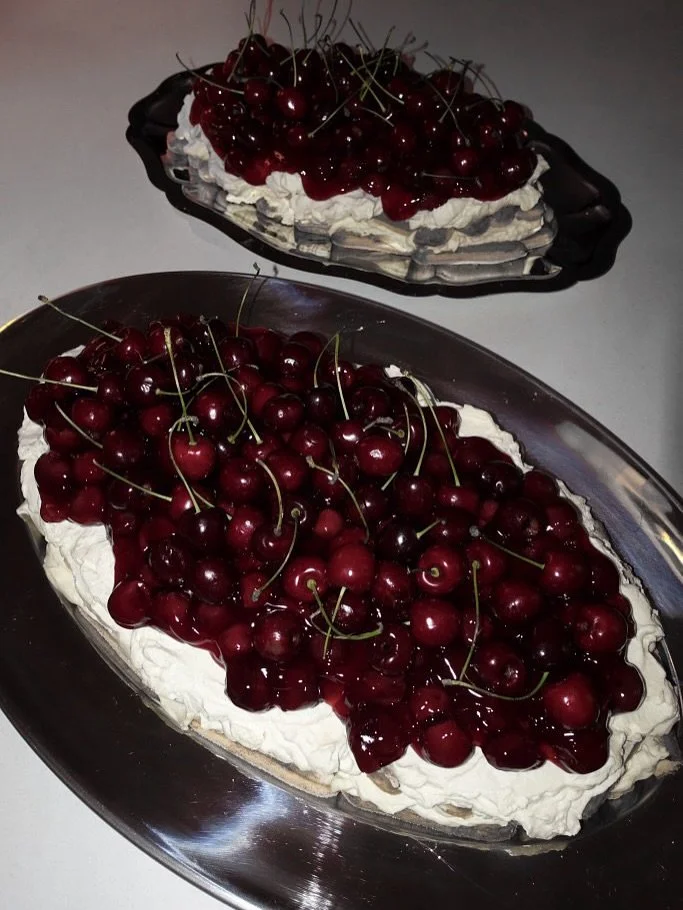 Een ovale chocoladetaart belegd met slagroom en verse kersen, gepresenteerd op een zilveren schaal, met een soortgelijke taart op de achtergrond.