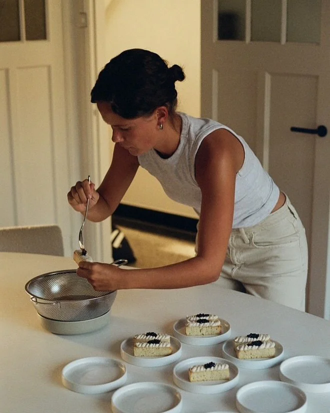 Iemand met kort donker haar en lichte huid, die over een tafel buigt en bezig is met eten opmaken. Op de tafel staan meerdere stukjes taart op kleine witte bordjes, met veel borden ernaast voor een catering.