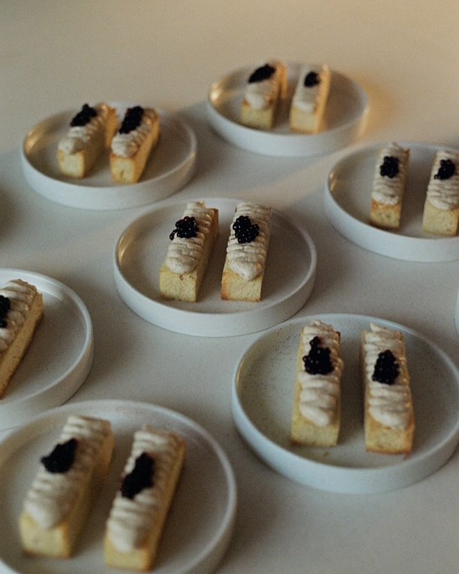 Meerdere kleine rechthoekige stukjes taart met witte frosting en zwarte bessen erop, gerangschikt op witte keramische bordjes voor catering.