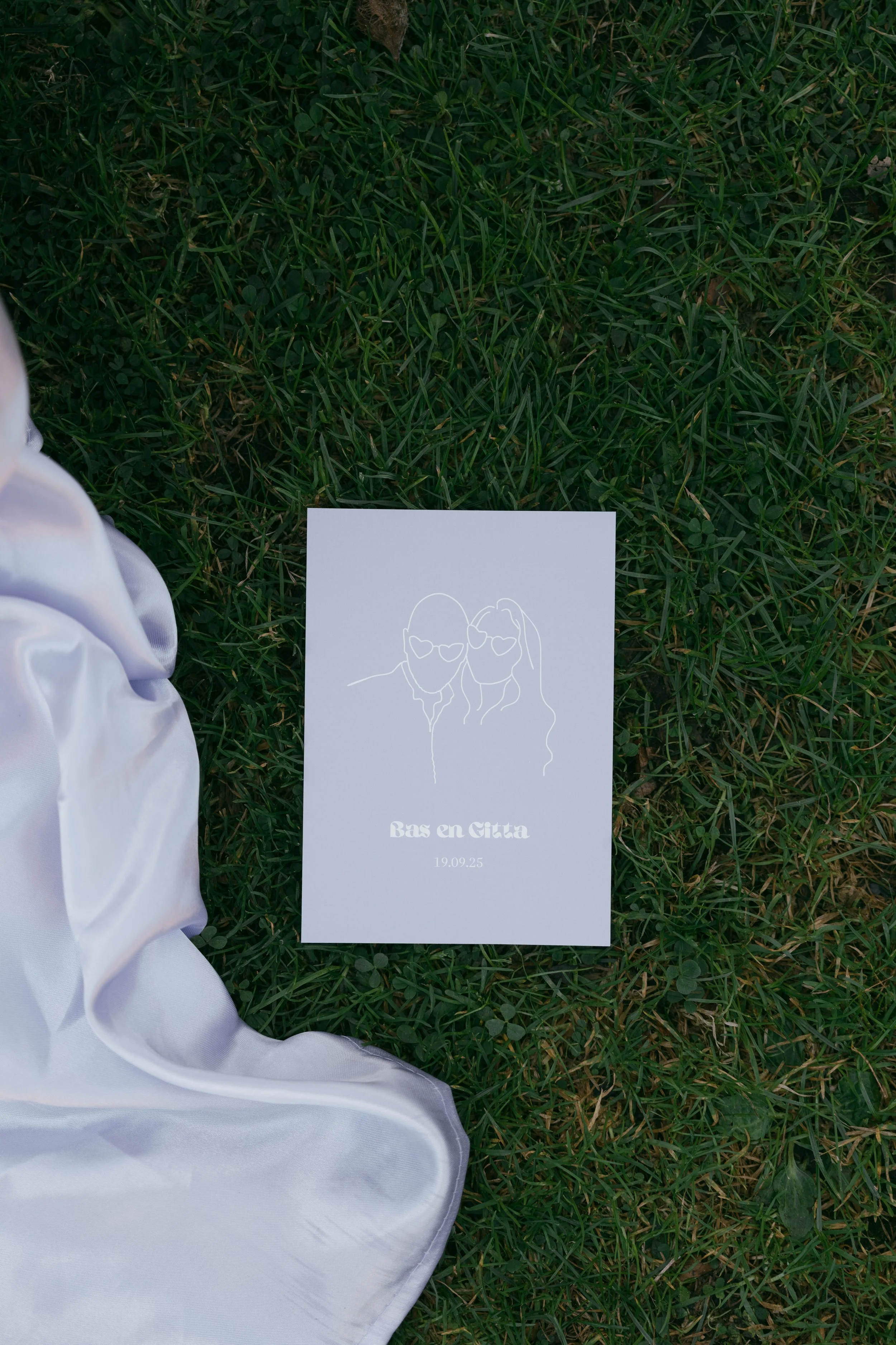 Lavendel lila paarse trouwkaart op het gras met een minimalistische lijntekening van een stel met zonnebrillen, samen met de tekst ‘Bas en Gitta’ en de trouwdatum. Aan de linkerkant is gedeeltelijk een satijnen witte doek zichtbaar.