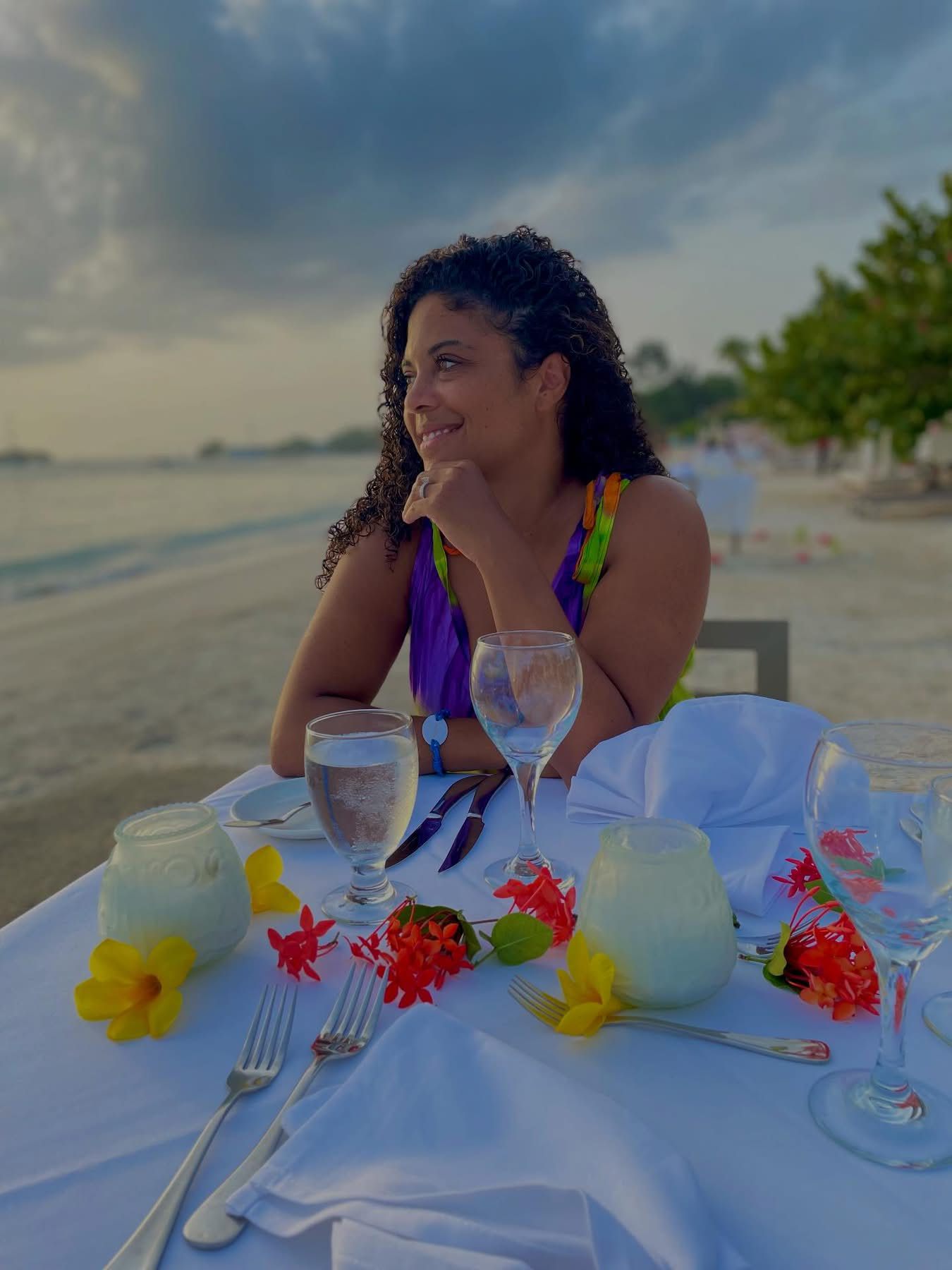 Author Hayden Fox sitting at a decorated outdoor beachside dining table in Jamaica with flowers, glasses, and cutlery, looking thoughtfully to the side with ocean and cloudy sky in the background.