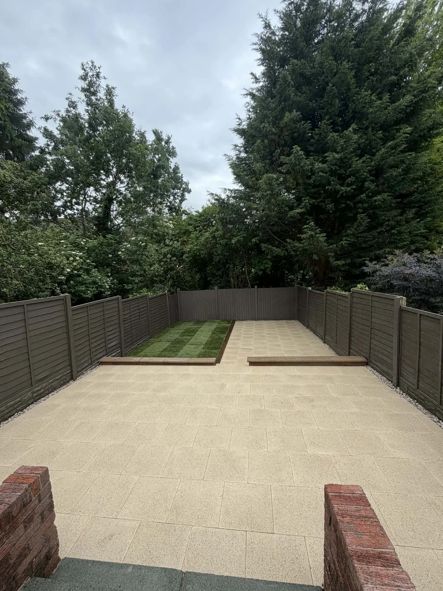 A backyard patio with beige paving, a green lawn patch, a gray weather fence, tall trees, and an overcast sky.