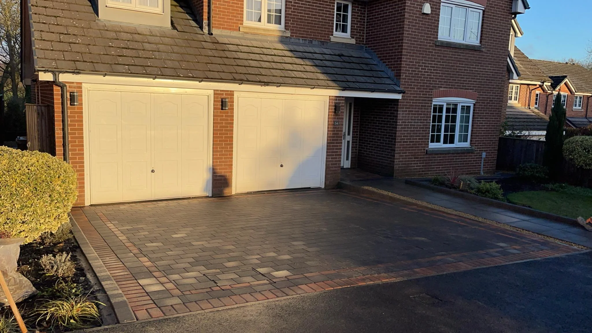 A modern brick house with a double garage door and a driveway made of brick pavers. There is a small garden with shrubs and a clear sky.