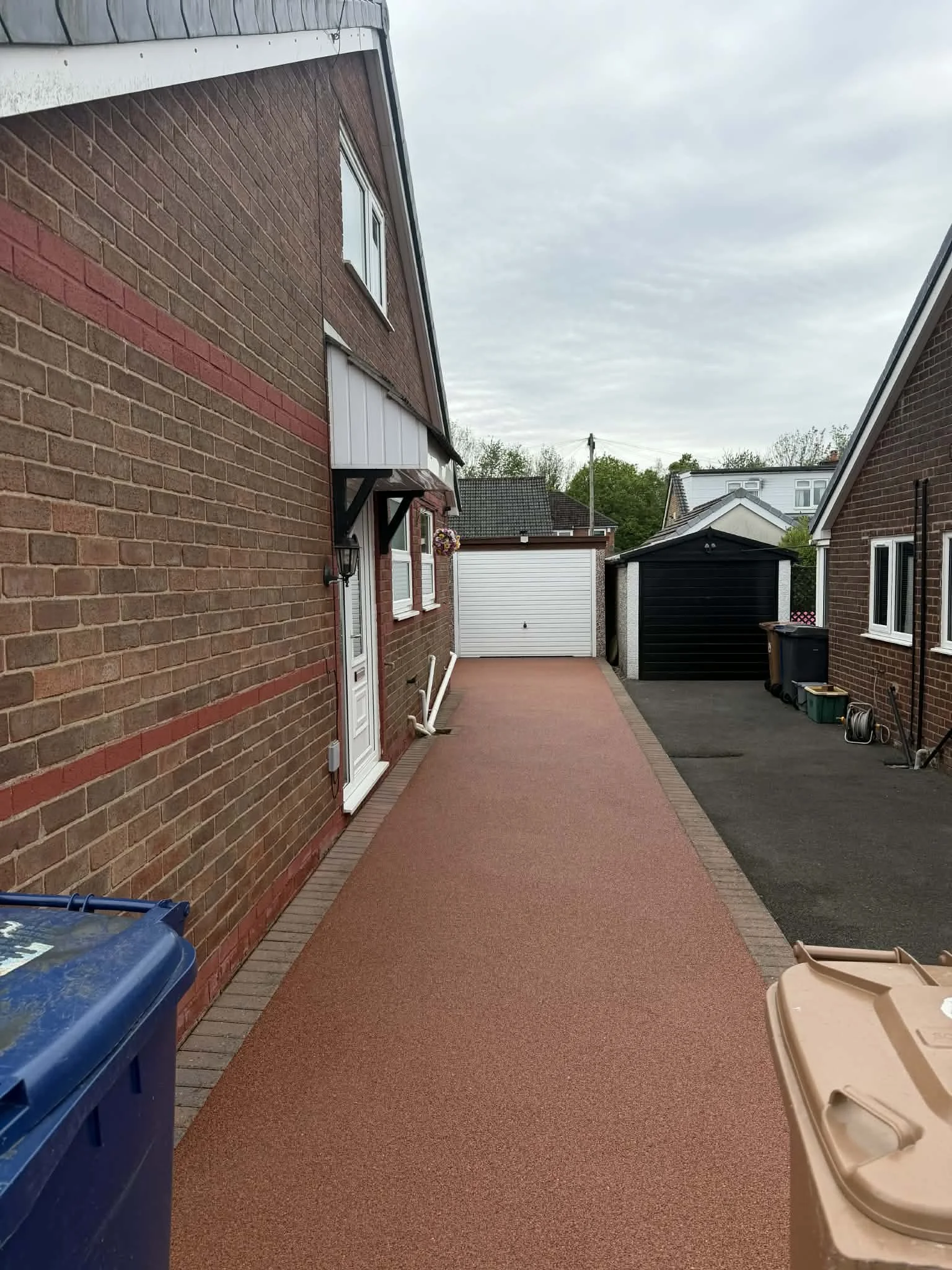 An alleyway between two brick houses with a red and black surface. There are garbage bins on the right and a blue recycling bin on the left. The sky is overcast.