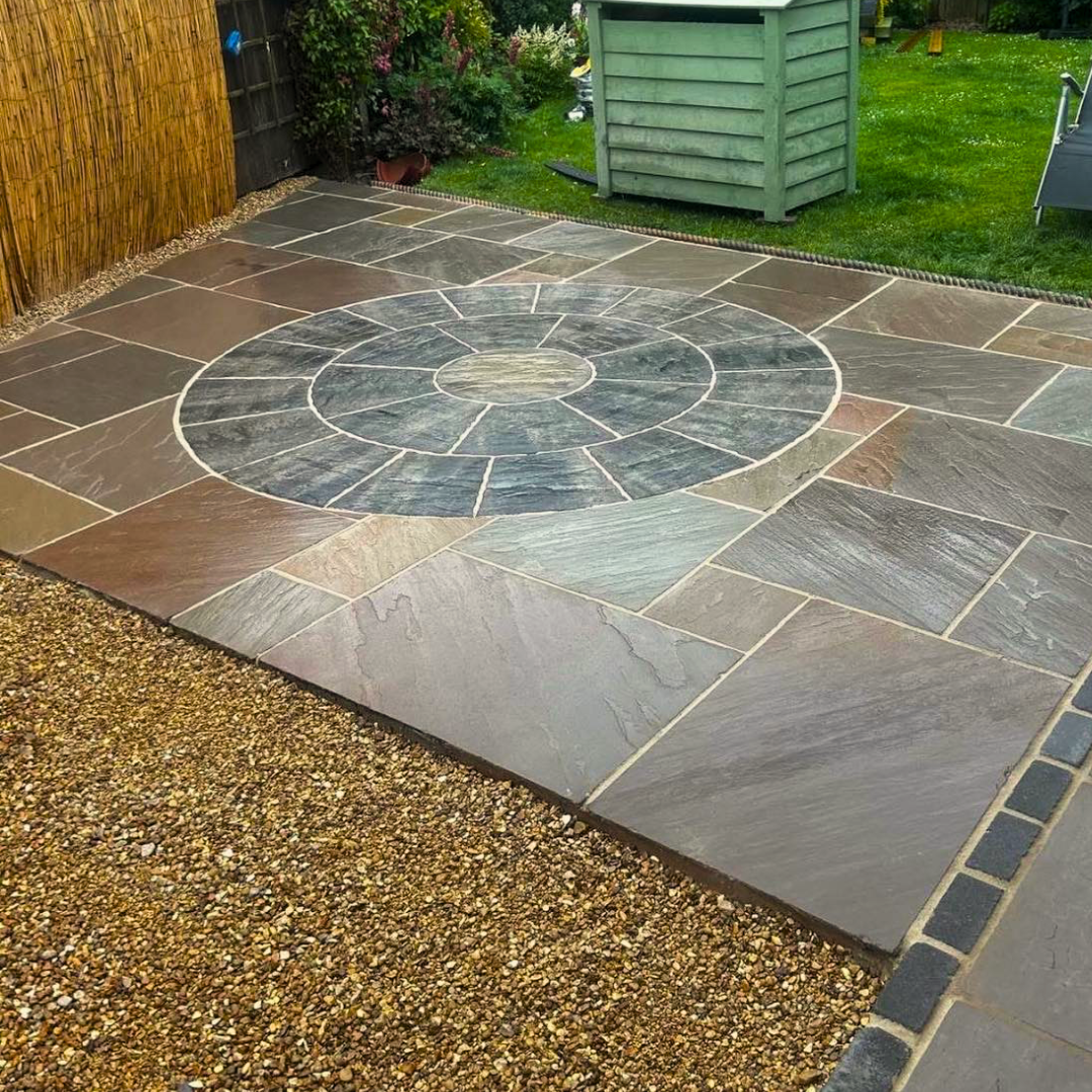 A backyard patio with a circular stone pattern in the center, surrounded by rectangular stone slabs, edged by gravel and a wooden fence on one side, with grass and a green shed in the background.