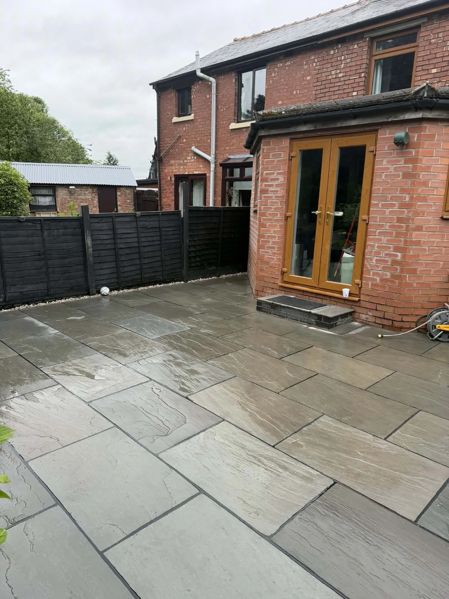 The image shows a backyard patio area with wet, gray stone tiles, a black wooden fence, and a brick house with wooden window frames and a set of French doors. There is a small step leading into the house, and a hose reel is visible on the right side.