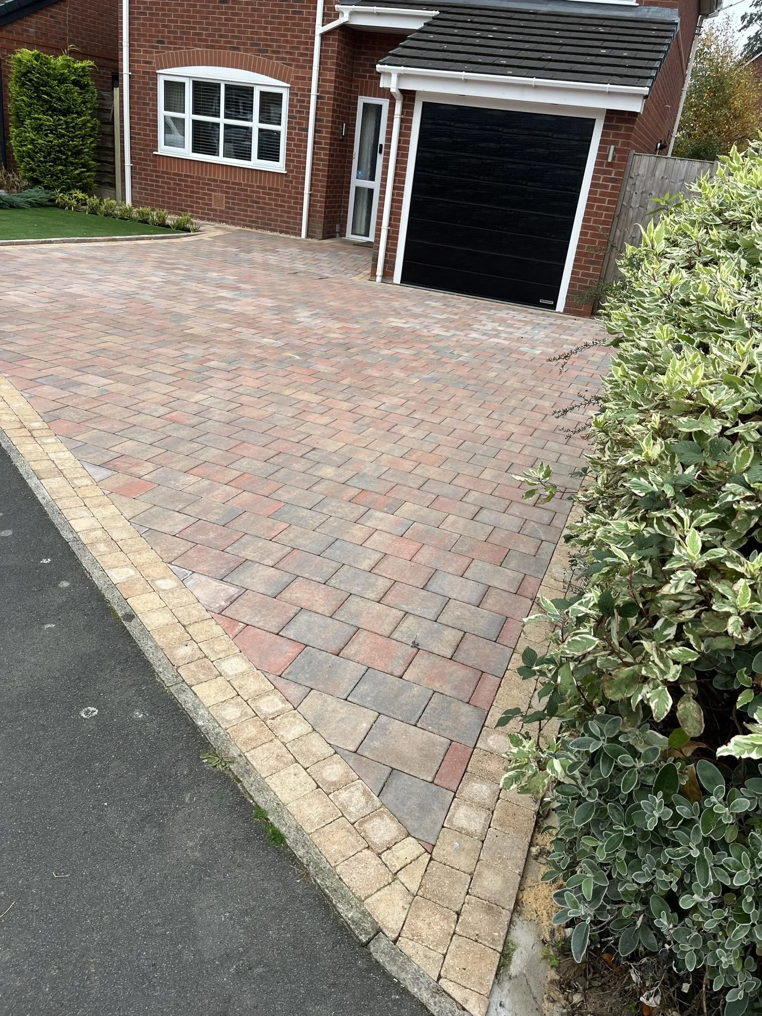 View of a paved driveway with brick pavers, a green shrub, and a brick house with white and black garage doors, a window, and some greenery on the side.