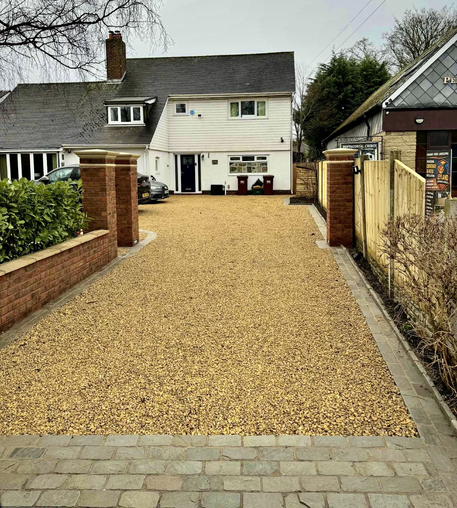 Front yard driveway of a house with a gravel surface, brick and wooden fencing, parked cars, and a house with white and beige exterior in the background.