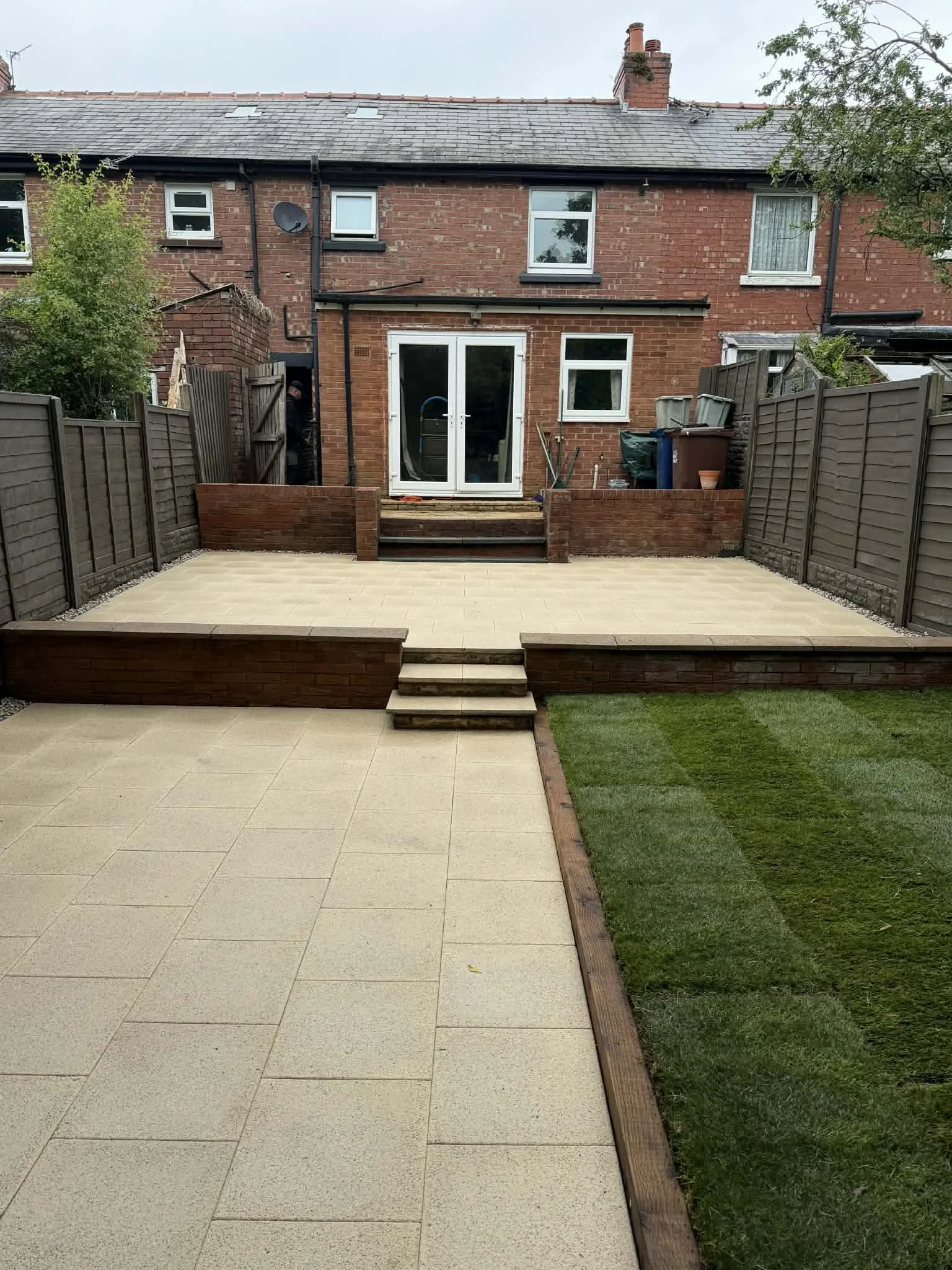 Backyard patio area with beige tiled flooring, a small set of stairs, and a garden with the lawn patch. The backyard is enclosed by brown wood fencing, with a brick house featuring windows and a glass door at the back. Some gardening tools and trash 