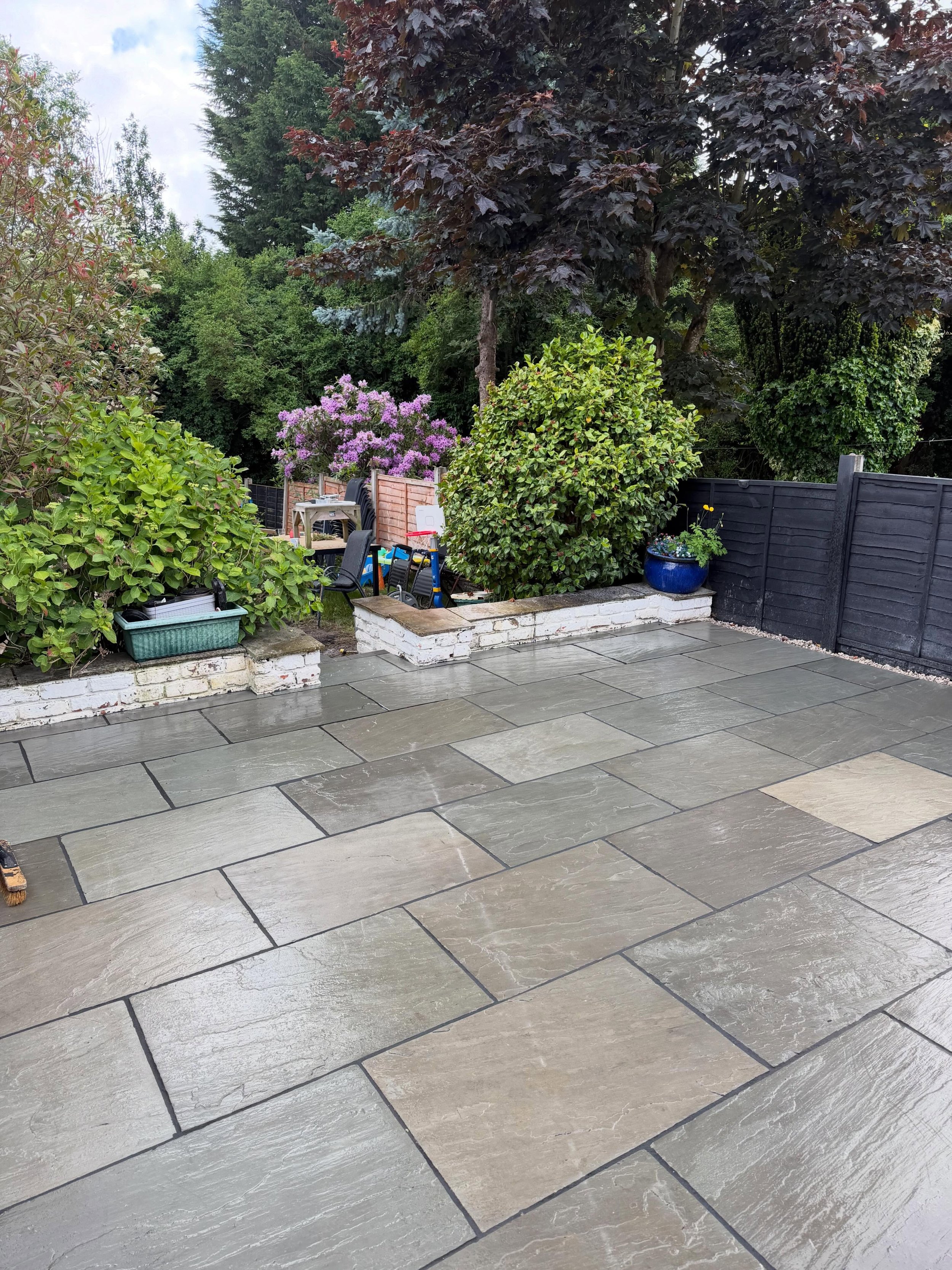 A backyard with a stone patio, various plants and shrubs, and a black fence. There are chairs, a small table, and gardening tools in the background.