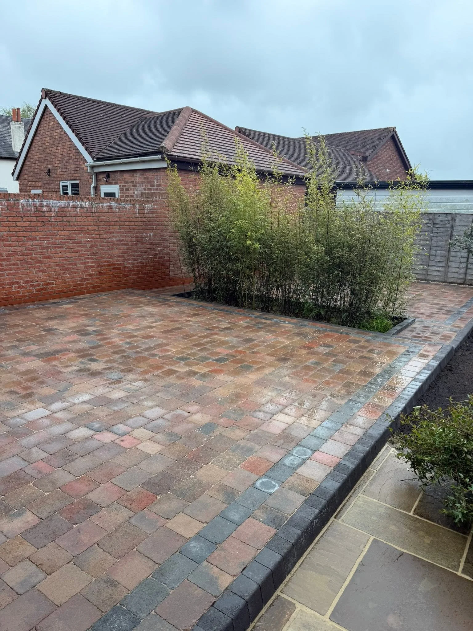 A paved backyard with brick and concrete border, brick wall, plants, and a brick house with a tiled roof and a cloudy sky.