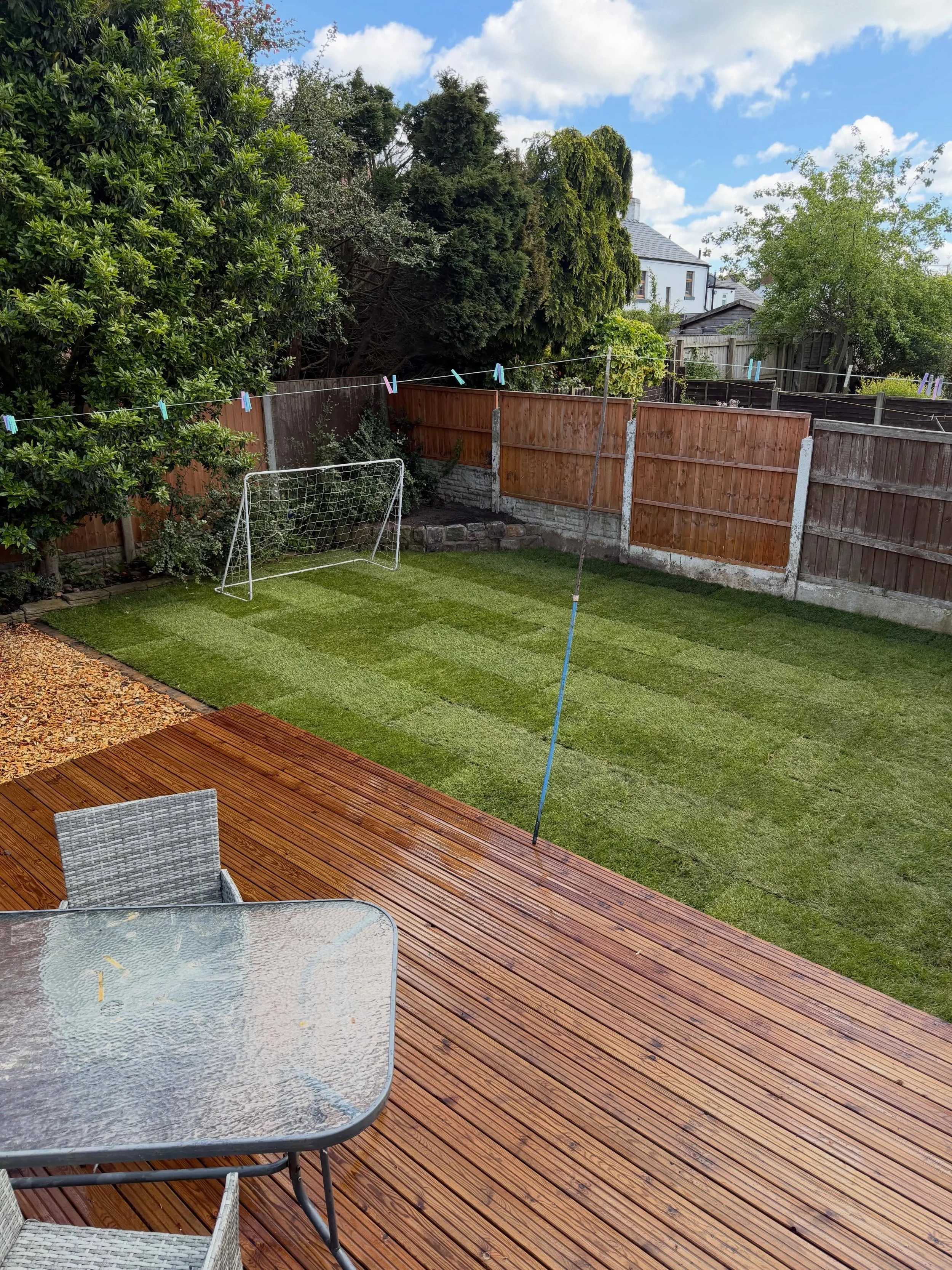 A backyard patio area with a glass-top outdoor table, two chairs, a wooden deck, and a fenced yard with a soccer goal, trees, and houses in the background under a partly cloudy sky.