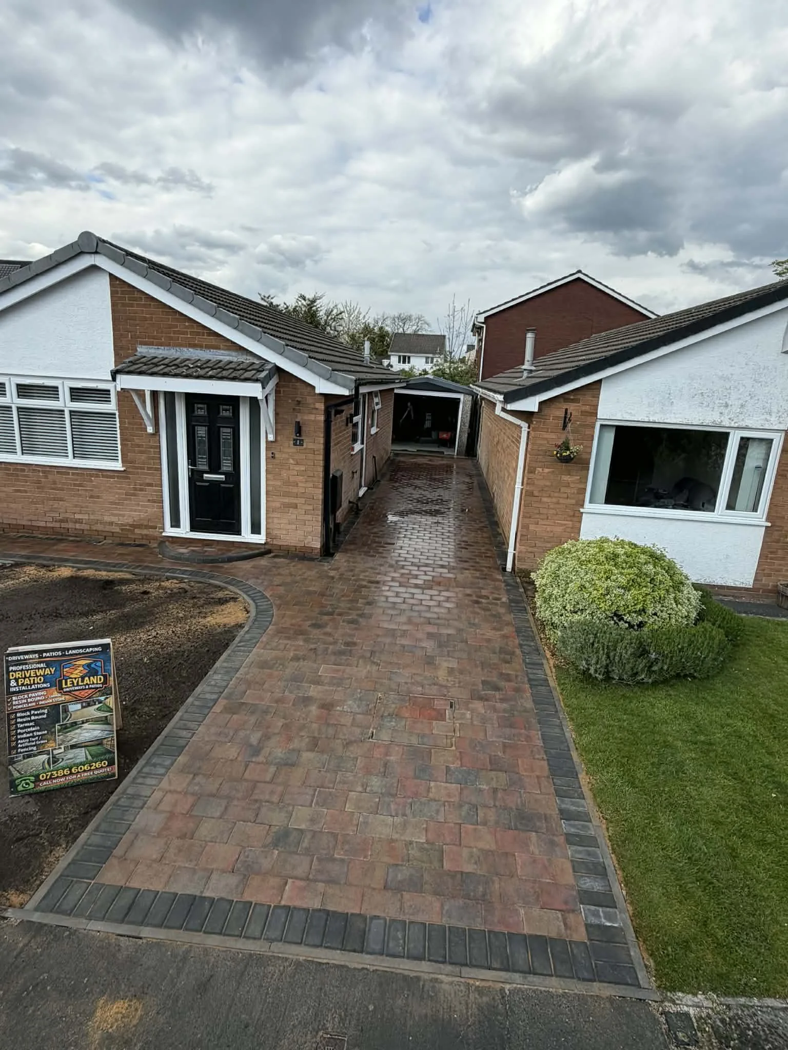 A paved driveway with brick and black border stones leading to a house with a black front door and large windows. To the left, a sign promotes driveway and patio installation services. The sky is cloudy.