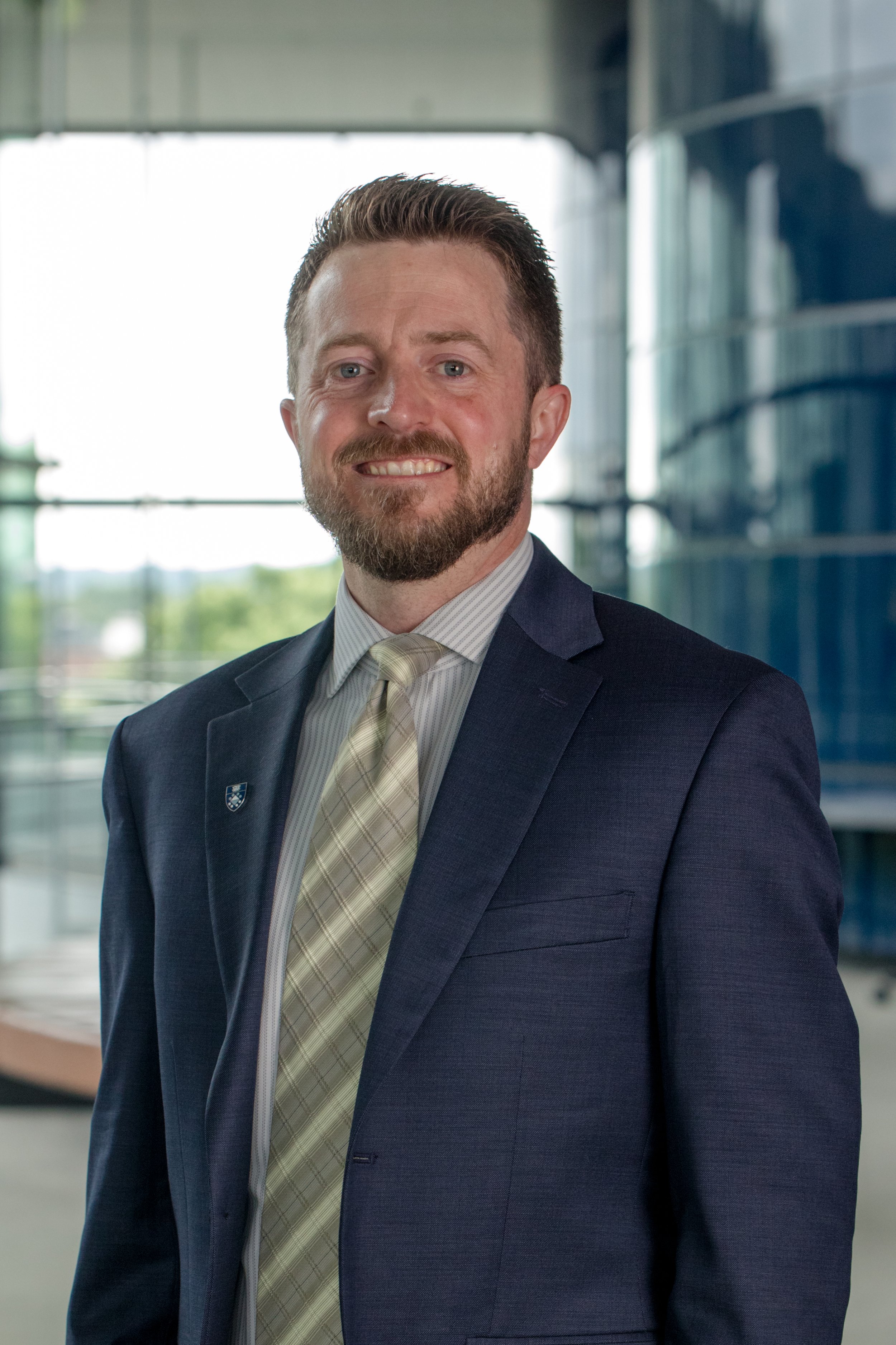Blake Christensen wearing a dark blue suit, light-colored tie, and white dress shirt, standing in front of a modern glass building.